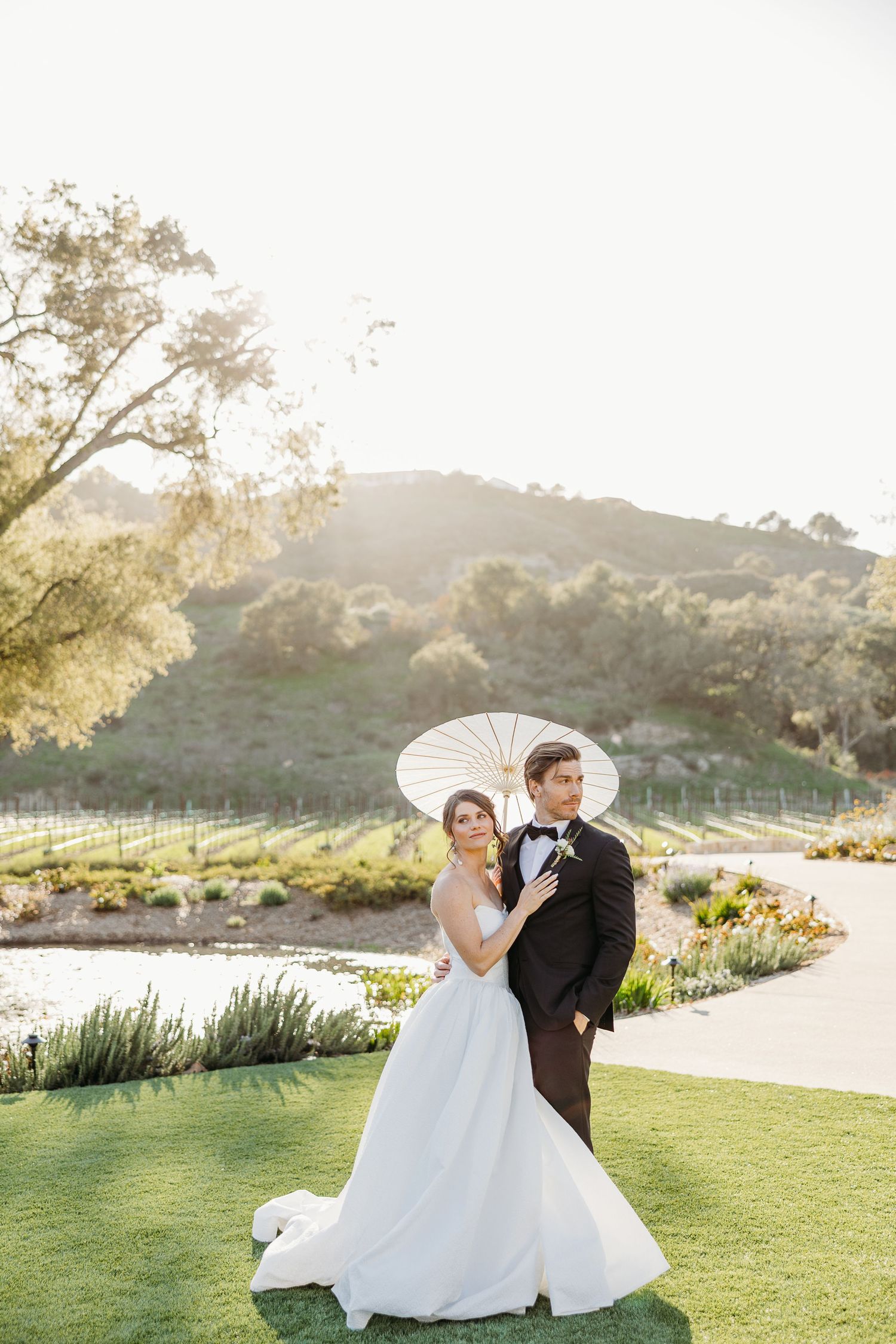 A couple in wedding attire poses together on a scenic grassy lawn overlooking a lake at sunset.