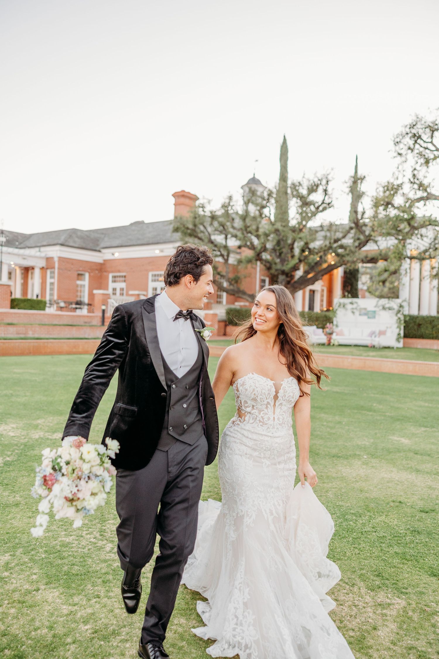 Newlyweds share a playful moment on a grassy lawn with classic brick buildings in the background.
