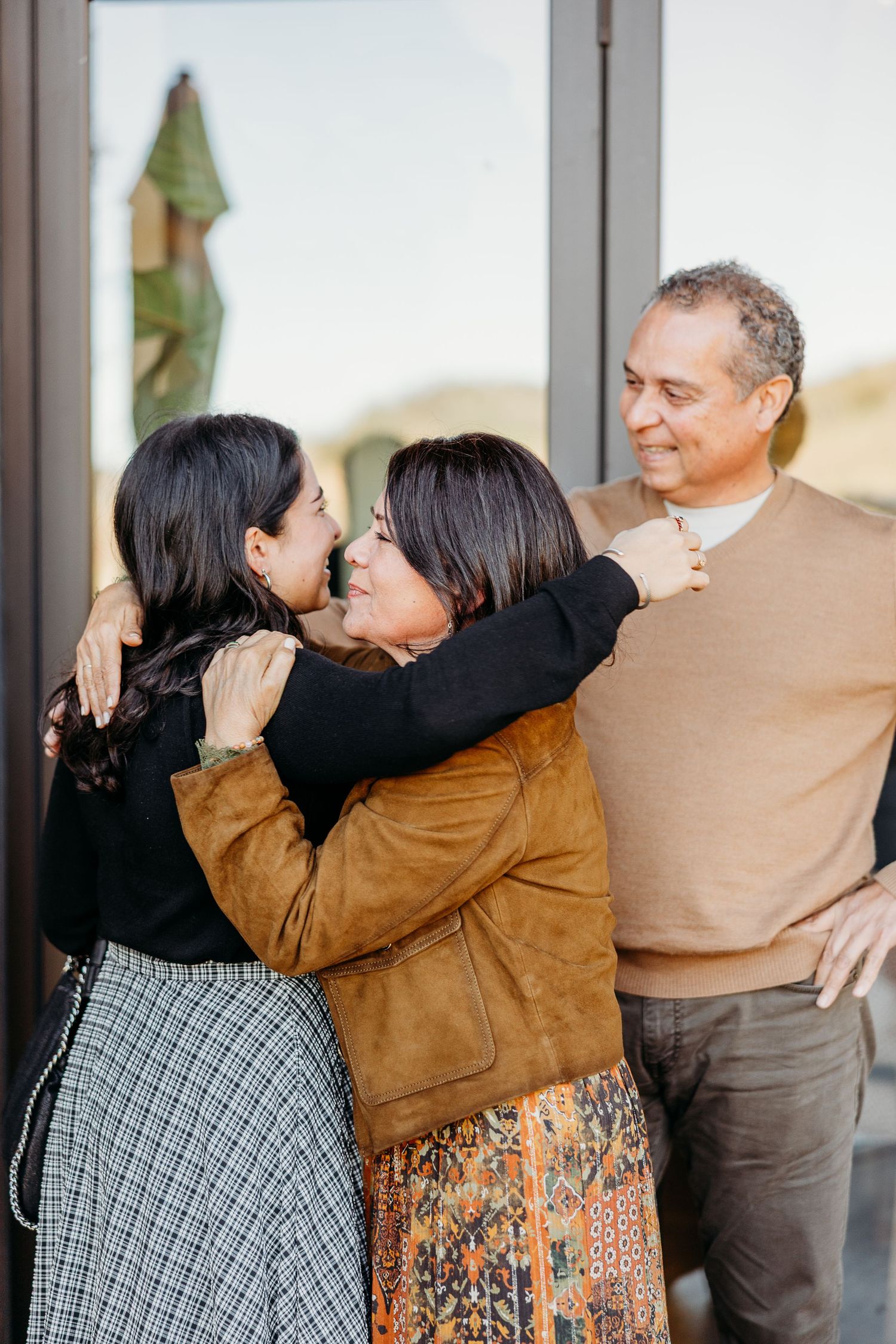 Three people embracing and sharing candid laughter against a light-colored wall background.