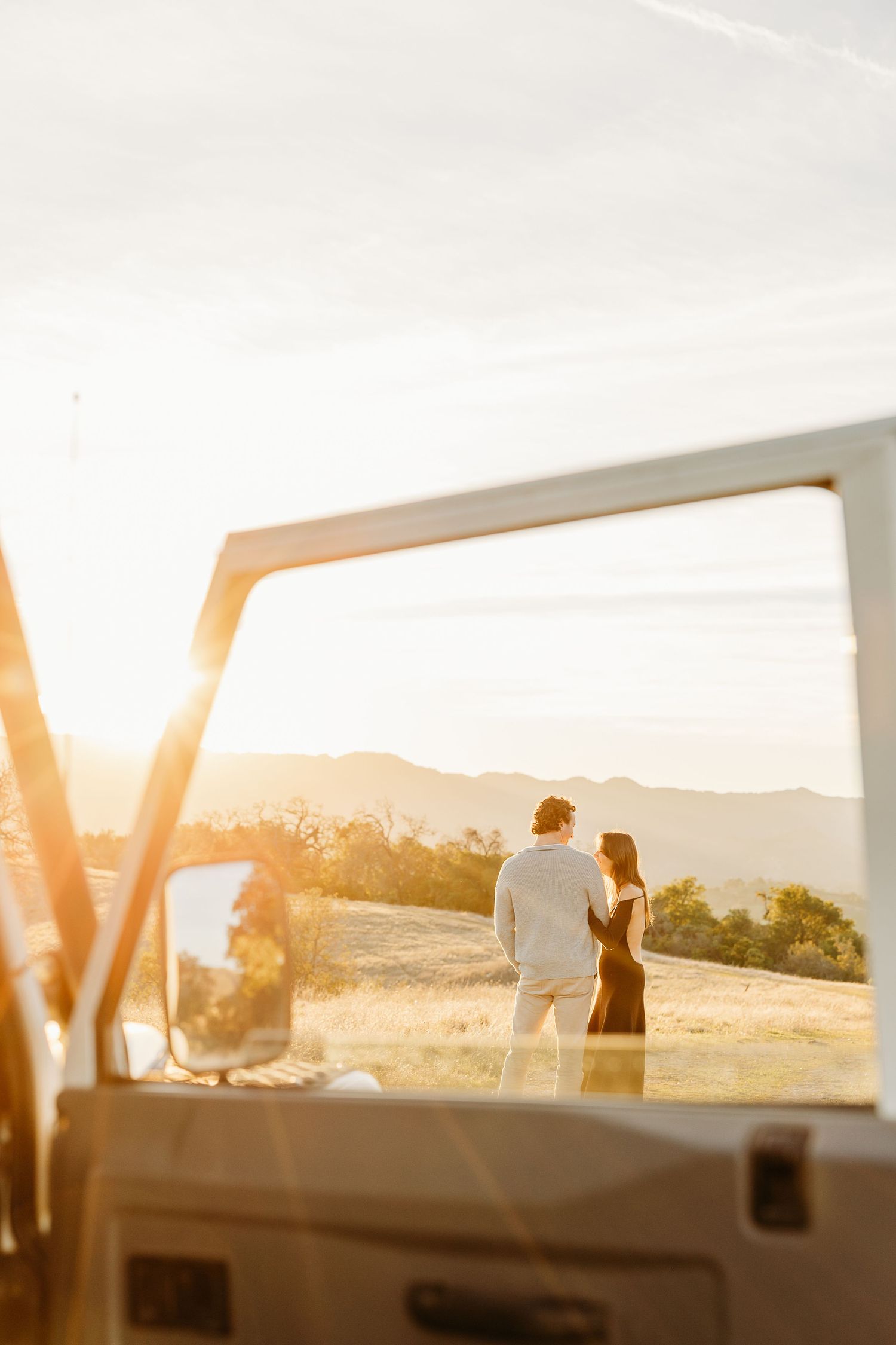 Sunset silhouettes through a car window frame with rolling hills in the background.