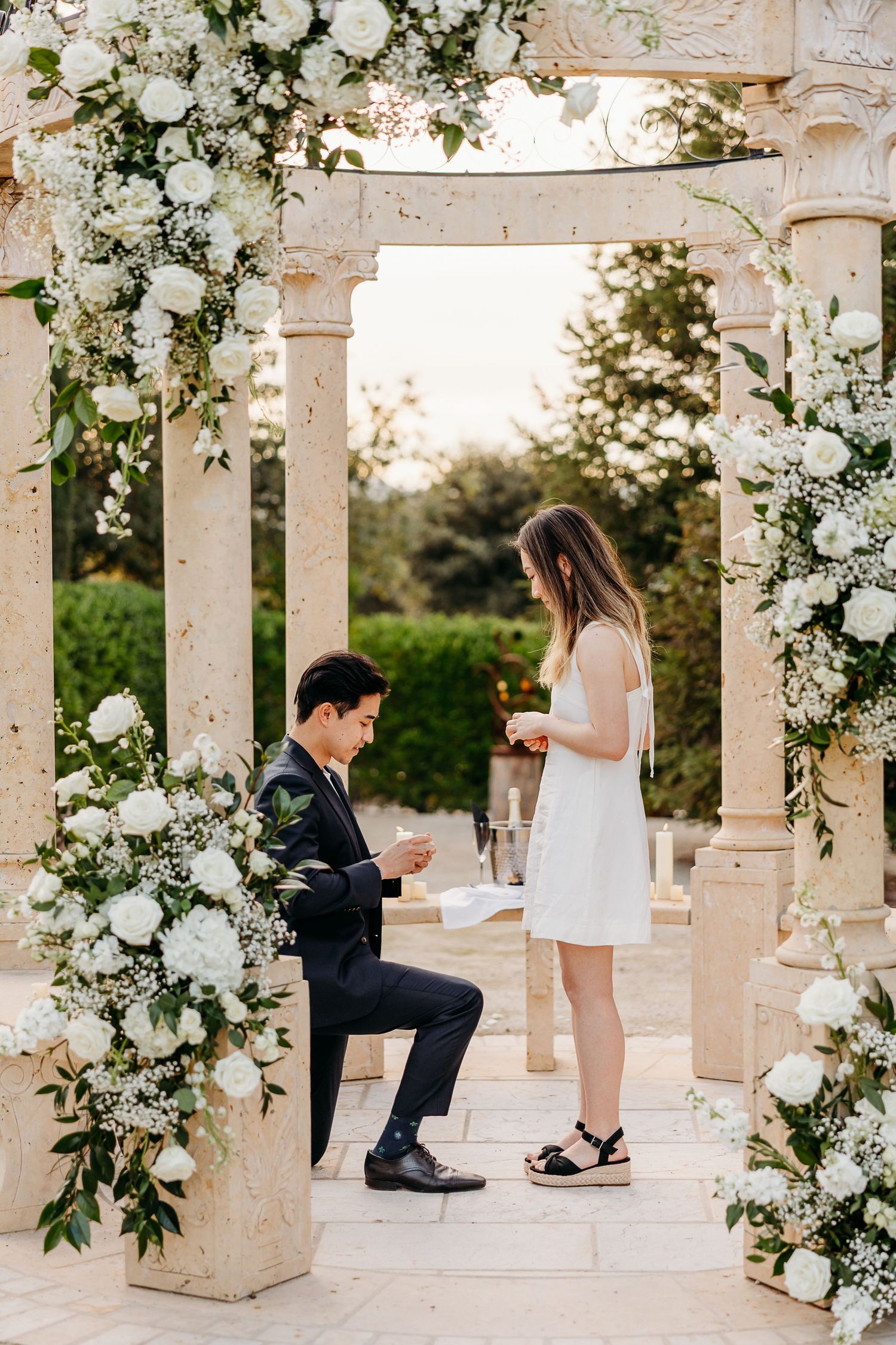 A romantic proposal scene under an elegant white floral arch at sunset in Allegretto Vineayrd Resort Garden.