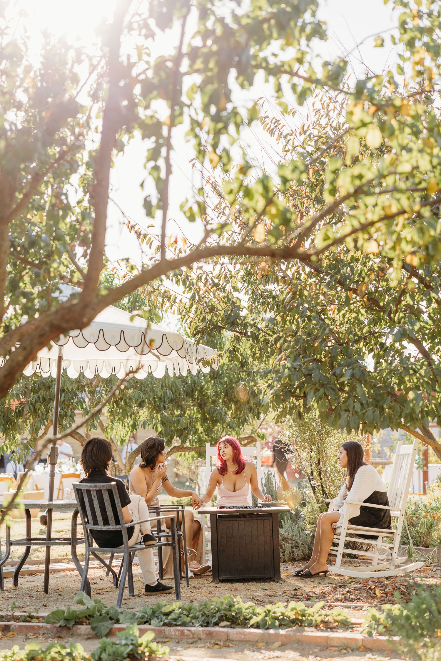 A group enjoys an outdoor gathering under a tree with string lights and a scalloped umbrella on a sunny afternoon.