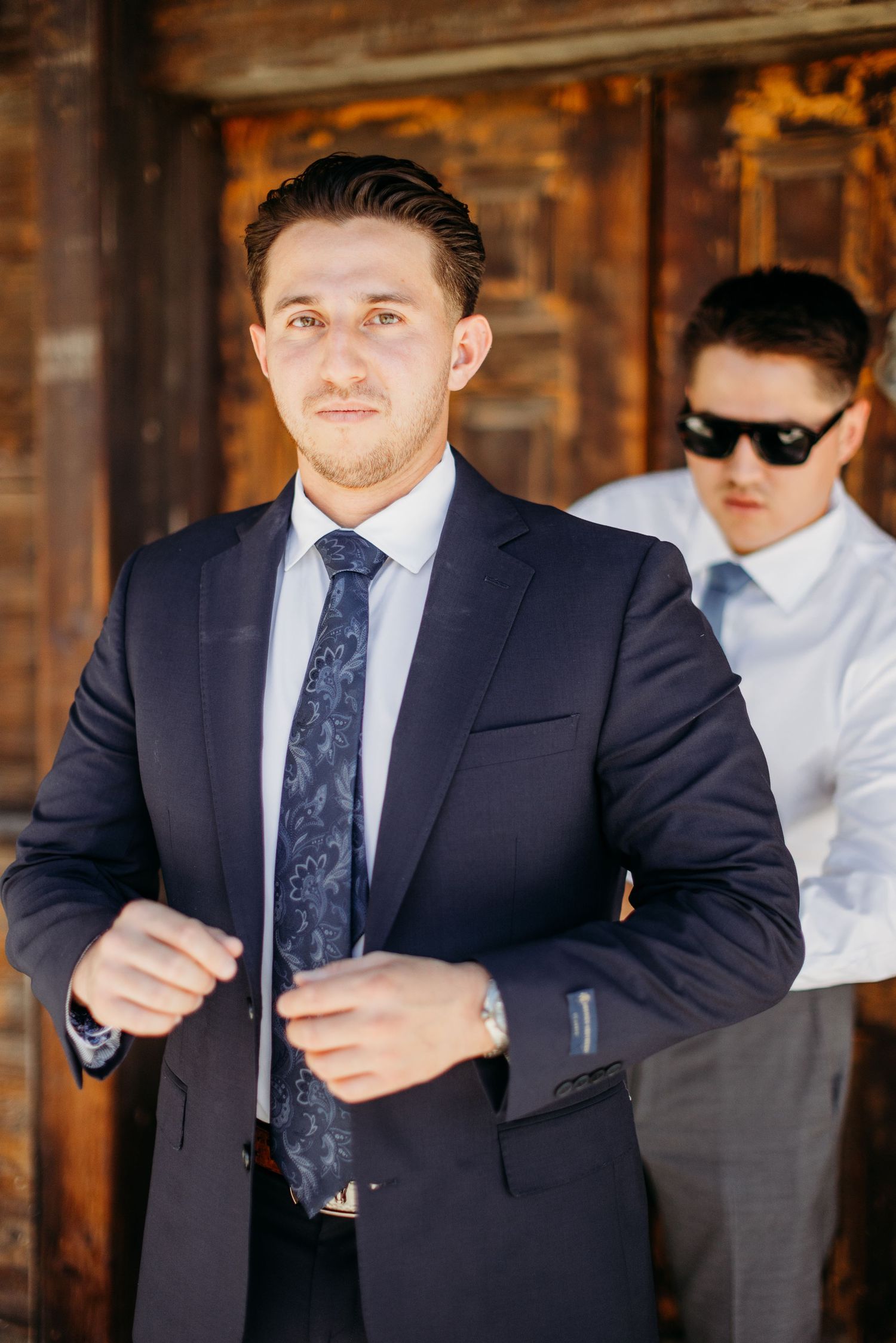 A person in a navy suit and tie adjusts their jacket while standing in a rustic wooden setting.