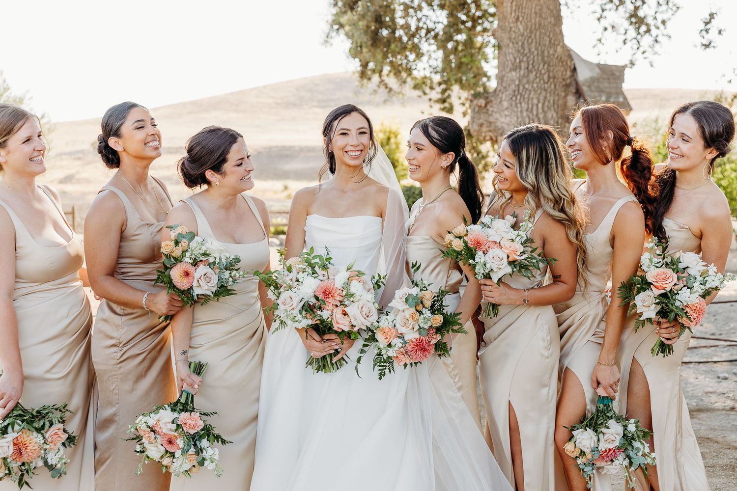 Bridesmaids in beige dresses laugh joyfully while holding coral and white floral bouquets at an outdoor wedding.