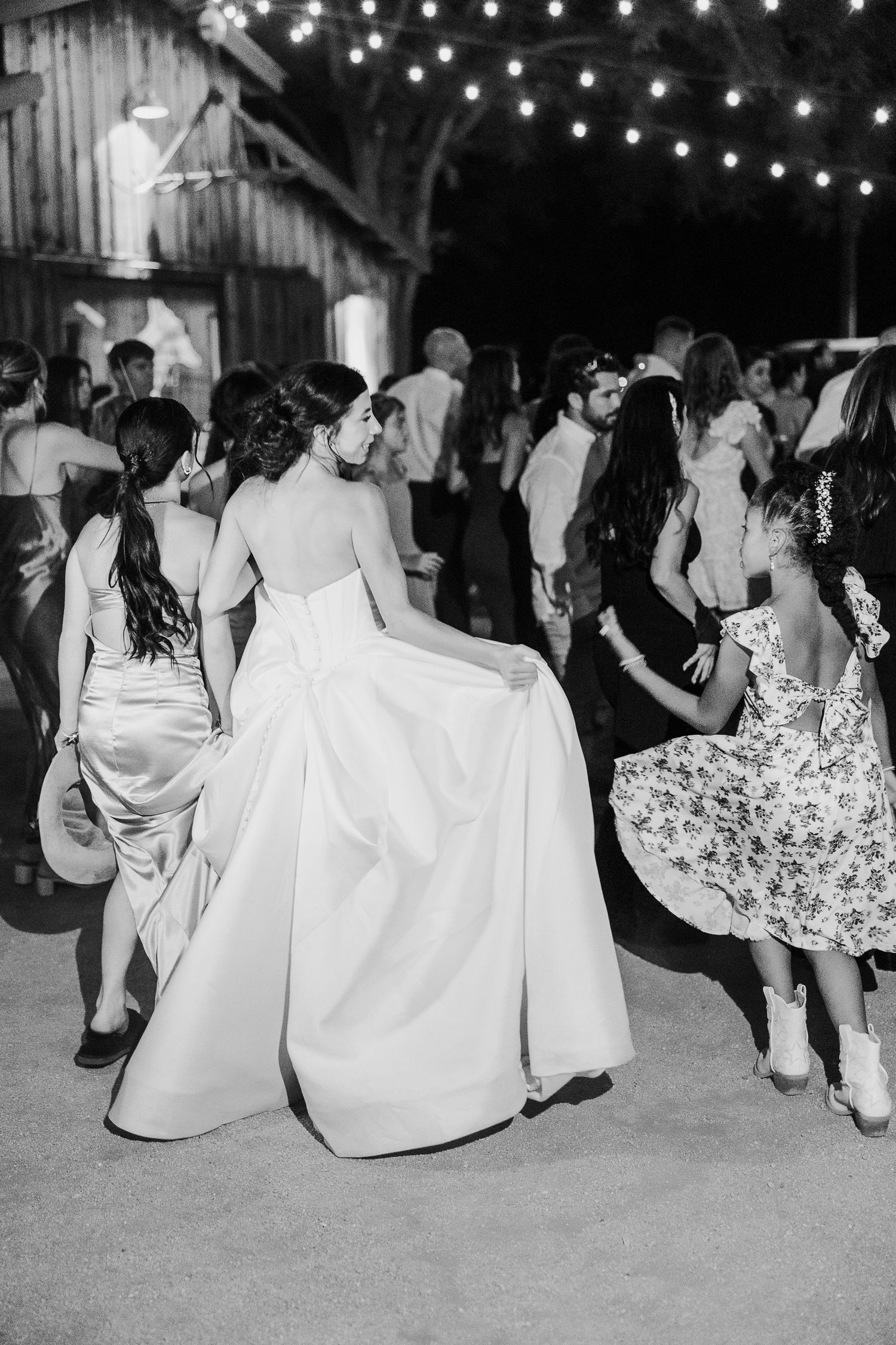 Black and white photo of wedding guests dancing under string lights at outdoor reception.