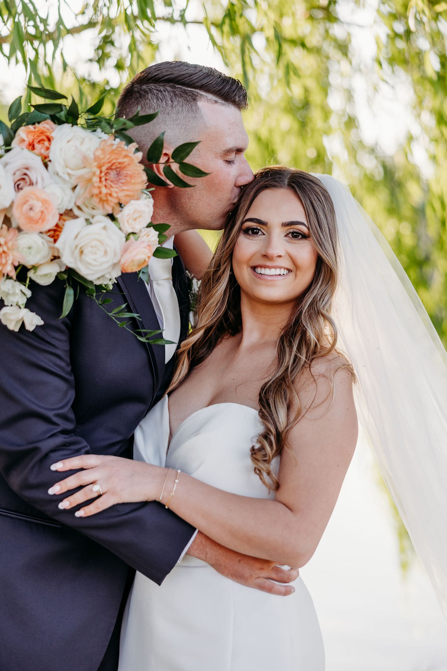 Romantic wedding photos show intimate moments between newlyweds against willow trees with peach and white floral bouquet.