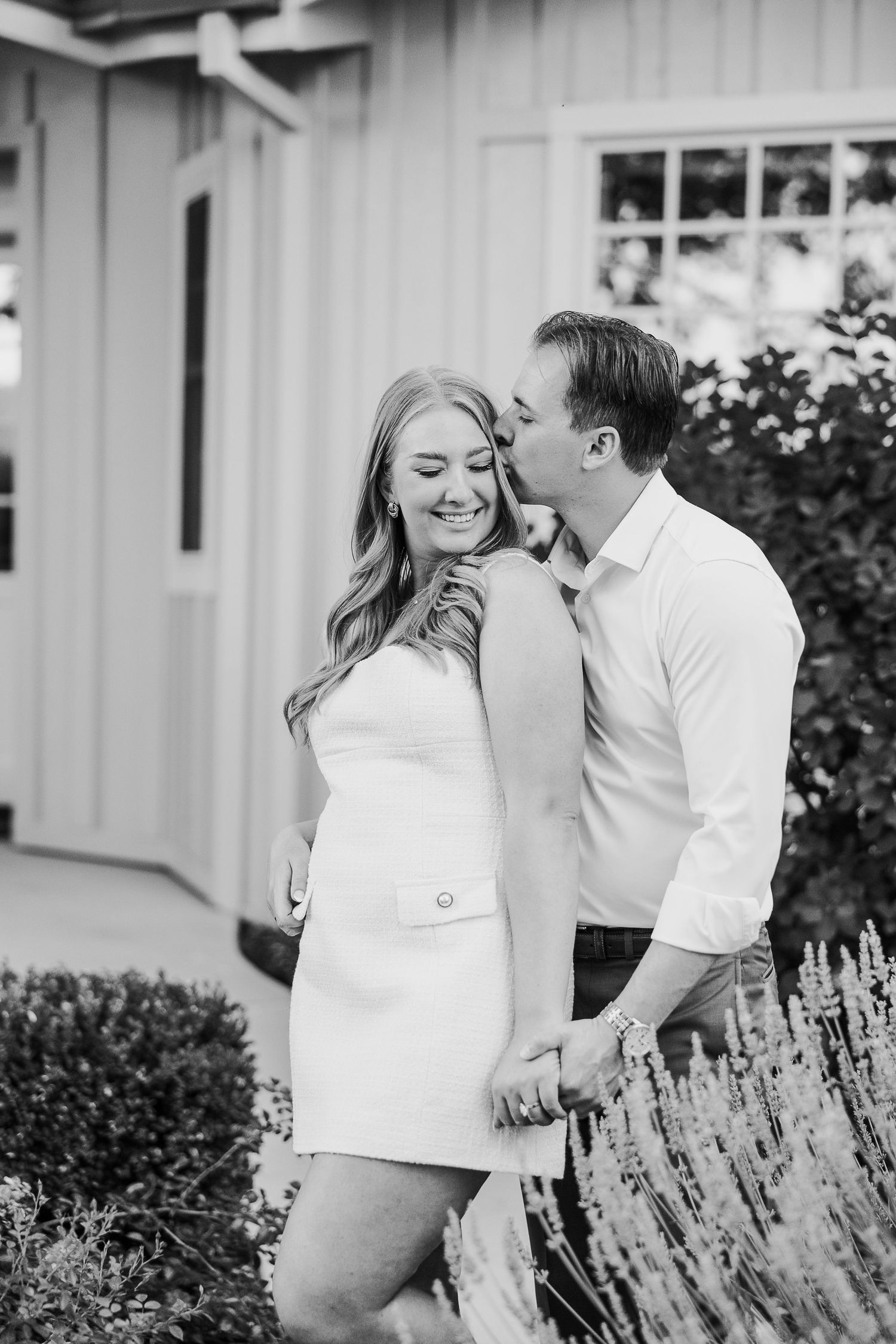 A romantic couple in white clothing poses together against a backdrop of lavender plants and a white building.
