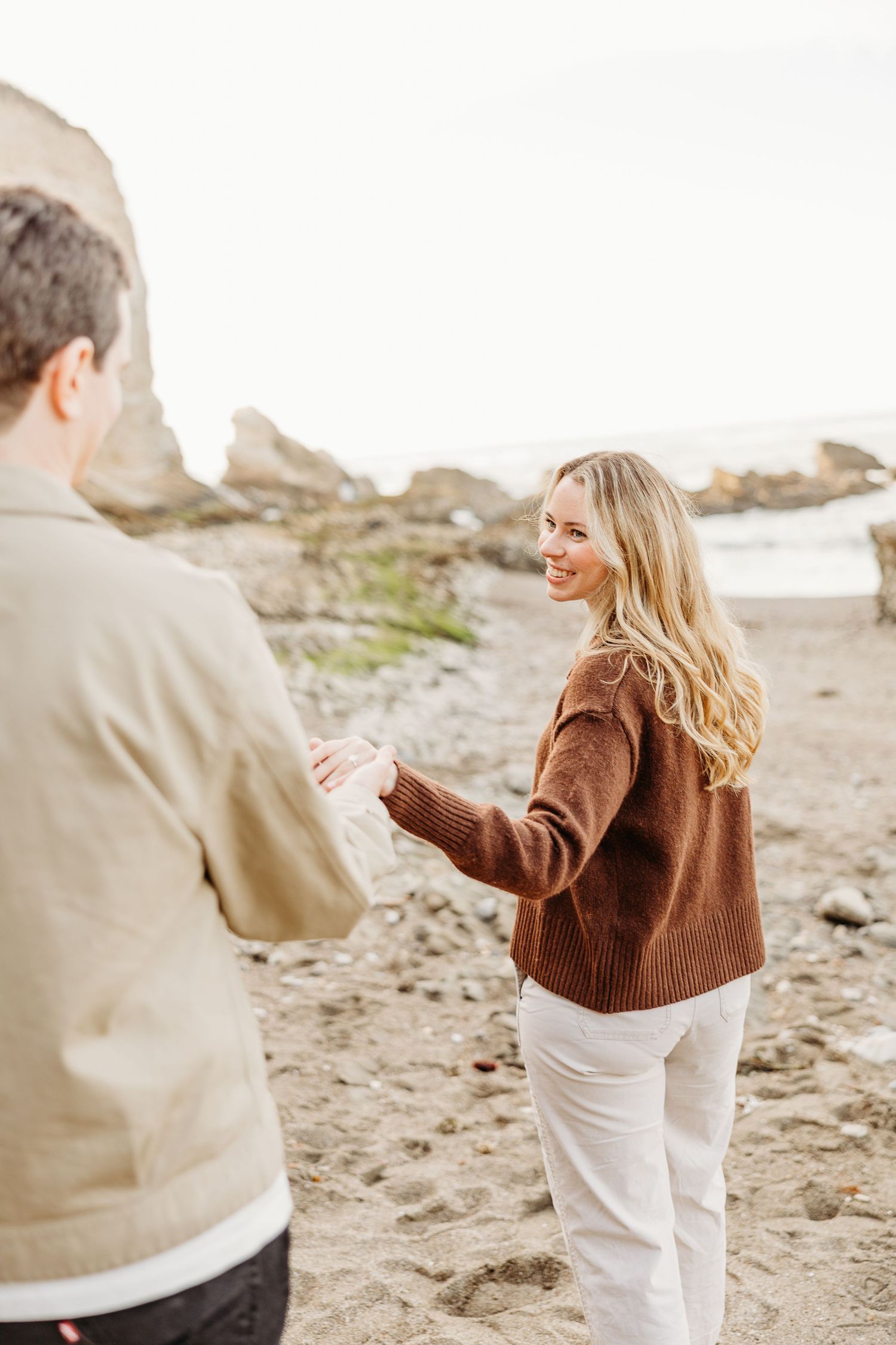 Playful couple in casual attire dancing and laughing together on a sandy beach.