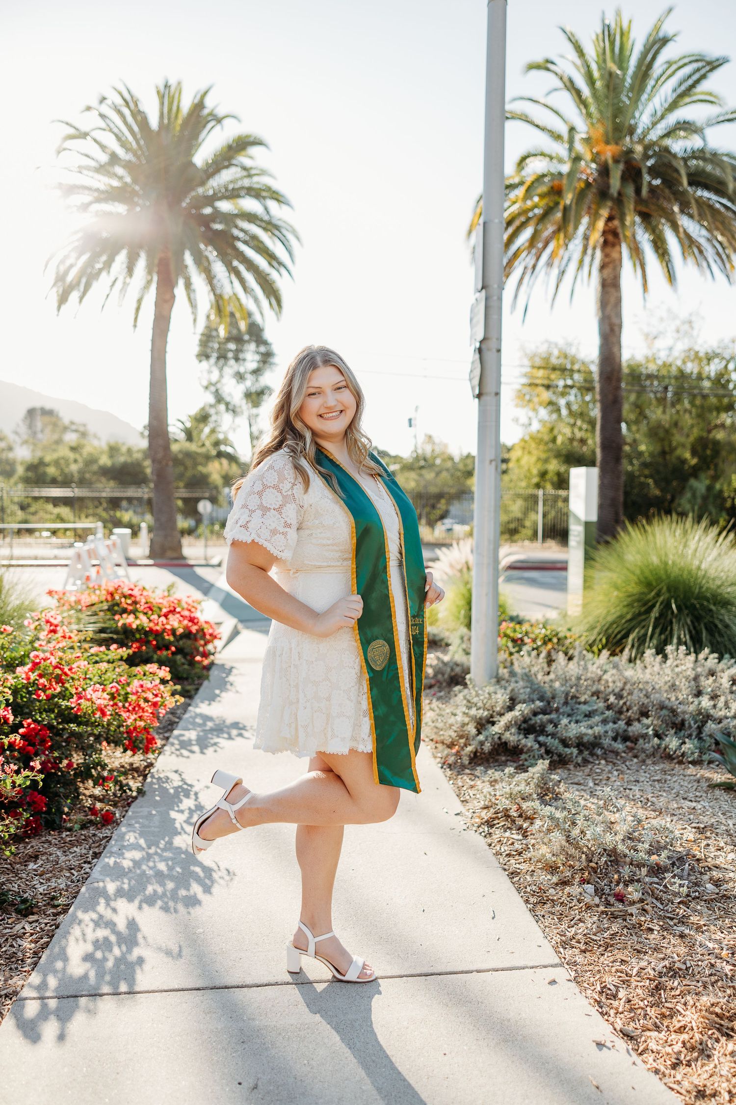 Someone in a white dress and green graduation stole poses on a sunny path with palm trees.