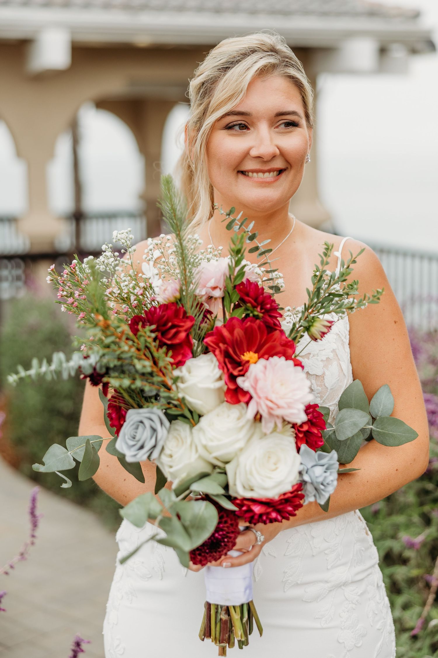 Joyful bridal portrait with vibrant red and white bouquet against Mediterranean architecture.