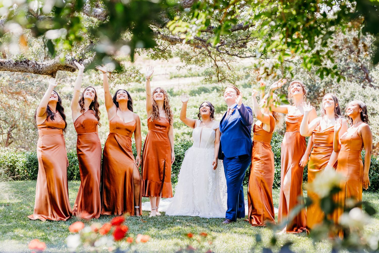 Wedding party in burnt orange dresses poses with couple outdoors.