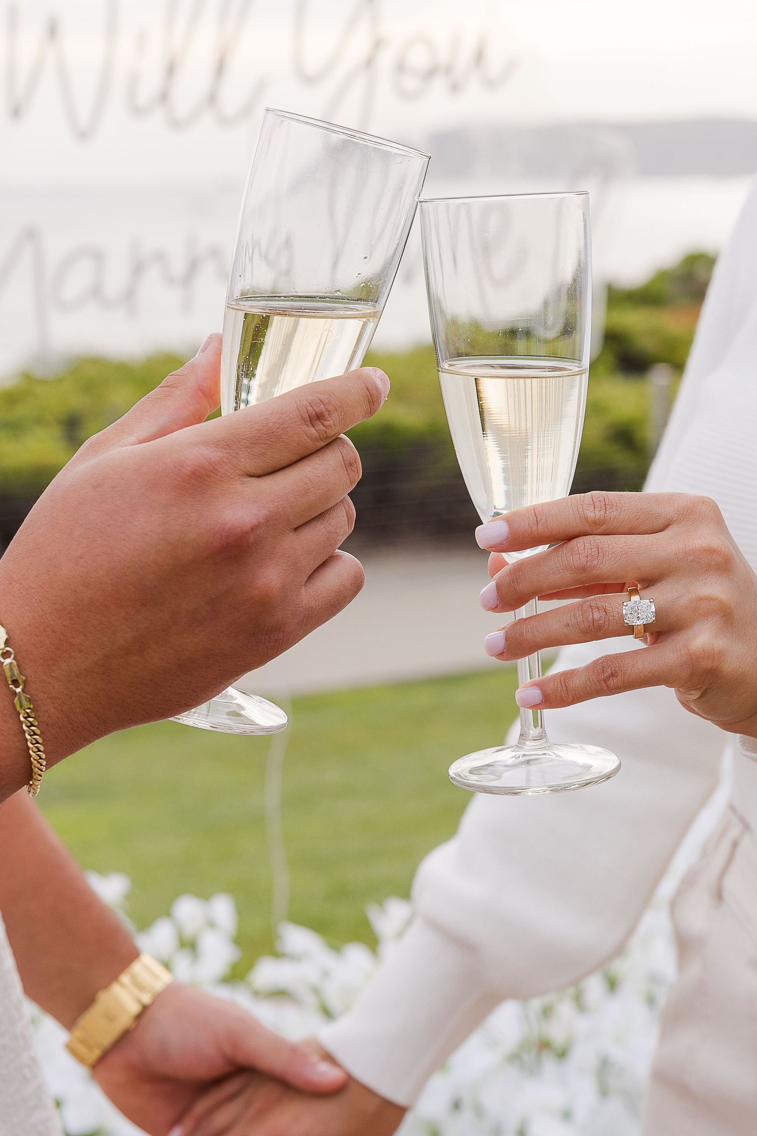Hands holding champagne glasses for a celebratory toast with engagement ring visible.