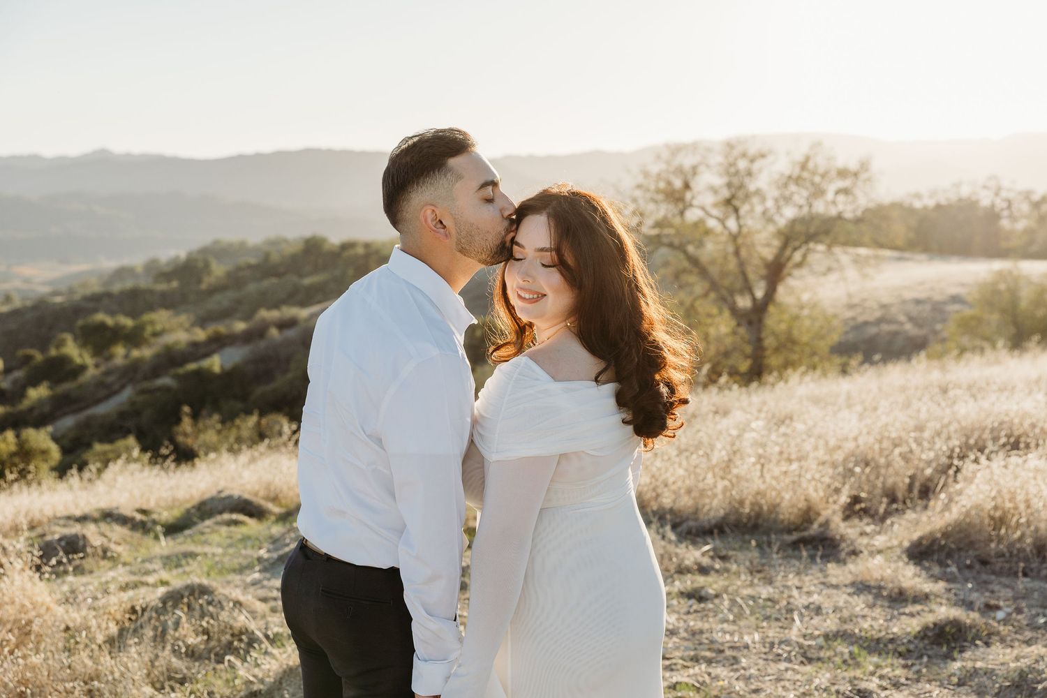 A romantic couple embraces in a sunlit meadow during golden hour as mountains create a dreamy backdrop.