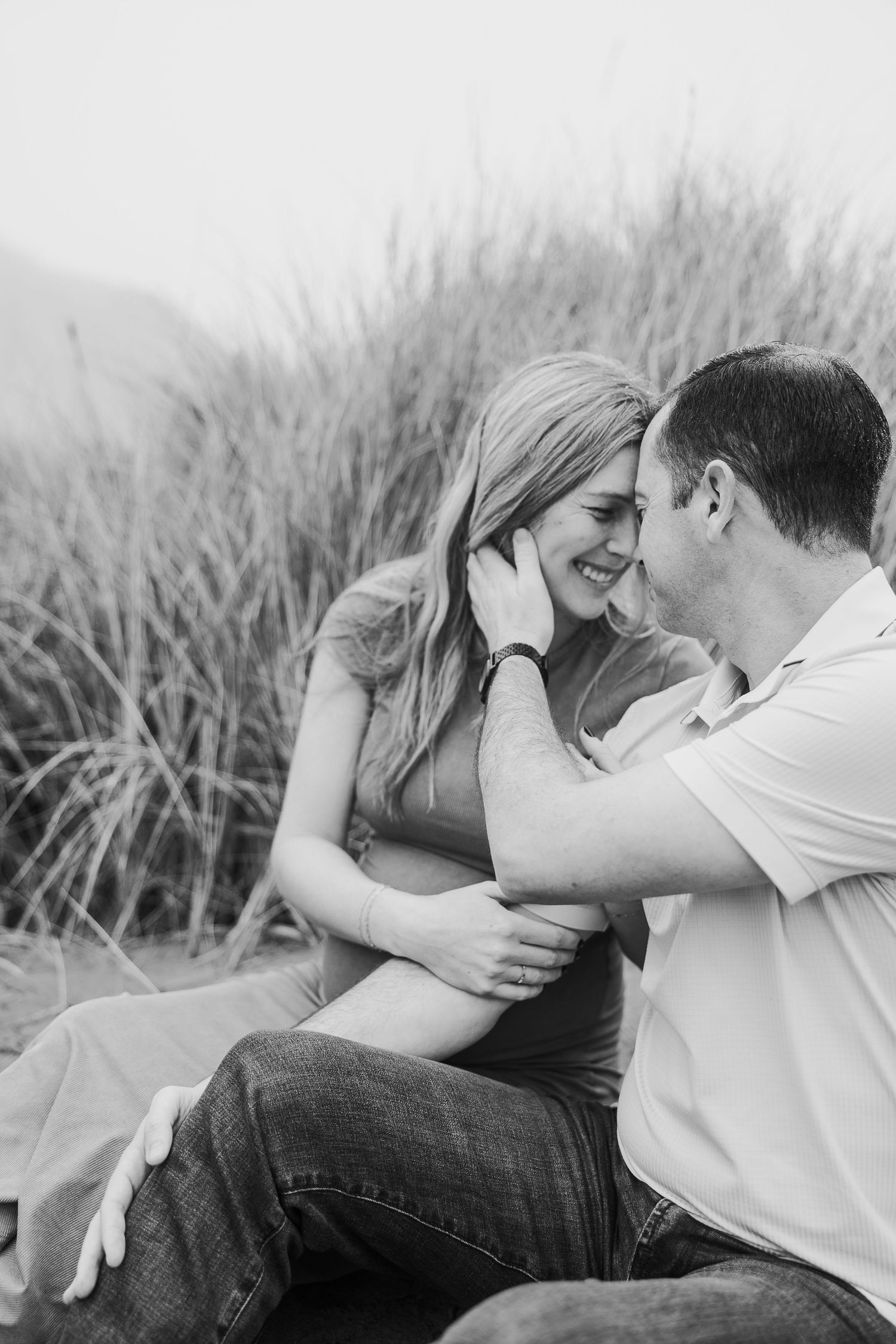 Black and white candid moment of couple sharing tender moment among coastal grass.