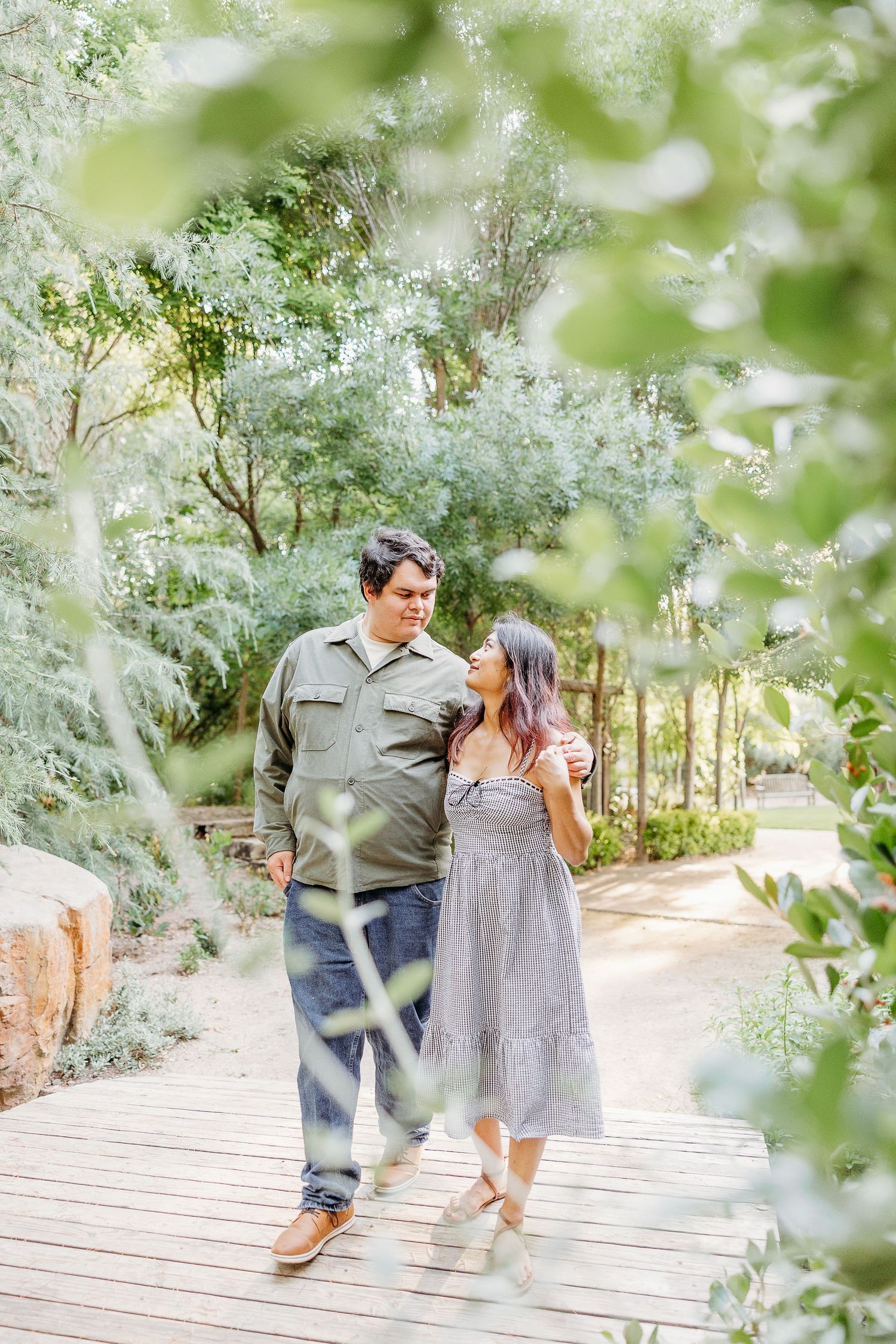 A couple walks together on a wooden pathway through a lush garden setting with soft natural lighting and greenery.