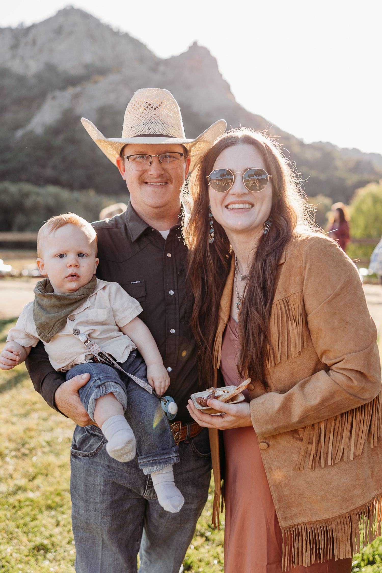 Western-styled family poses together outdoors with mountains in background at sunset.