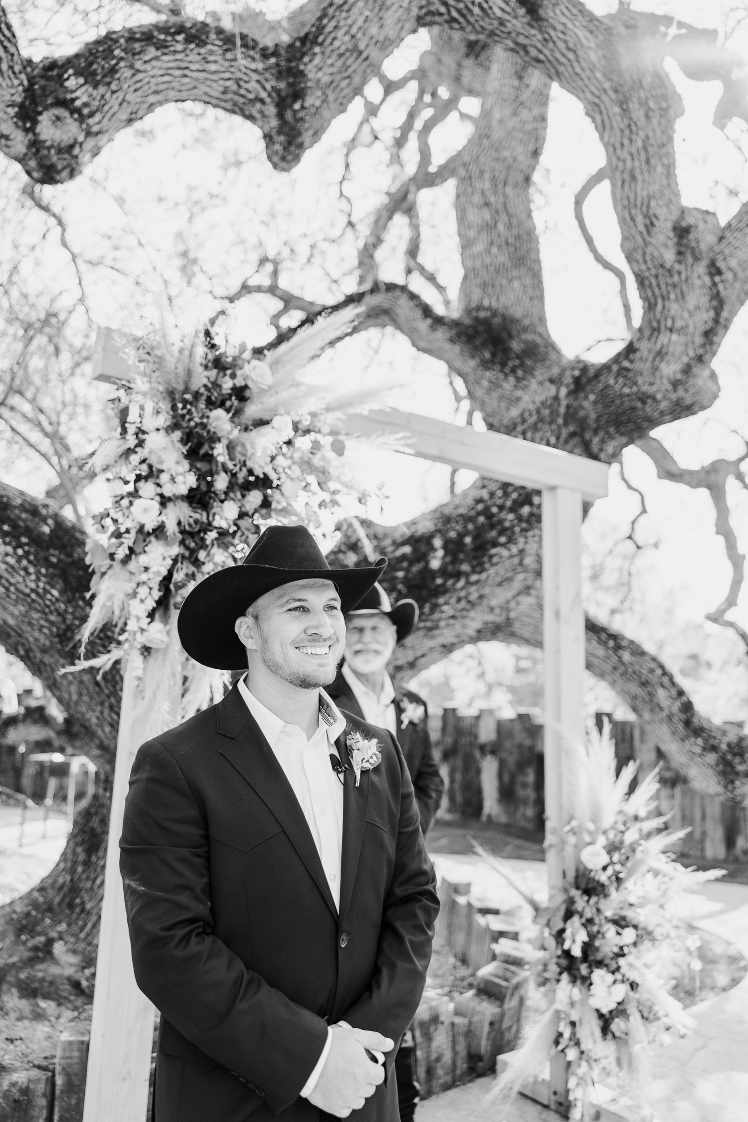 A groom in a black suit and cowboy hat smiles during an outdoor ceremony.