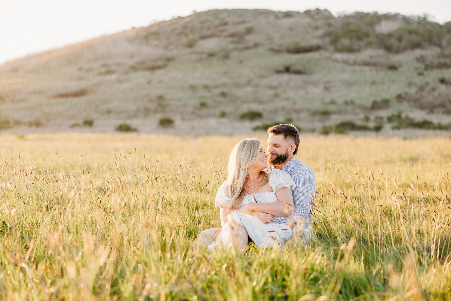 A romantic couple sits together in a sunlit golden wheat field at sunset with rolling hills in the background.