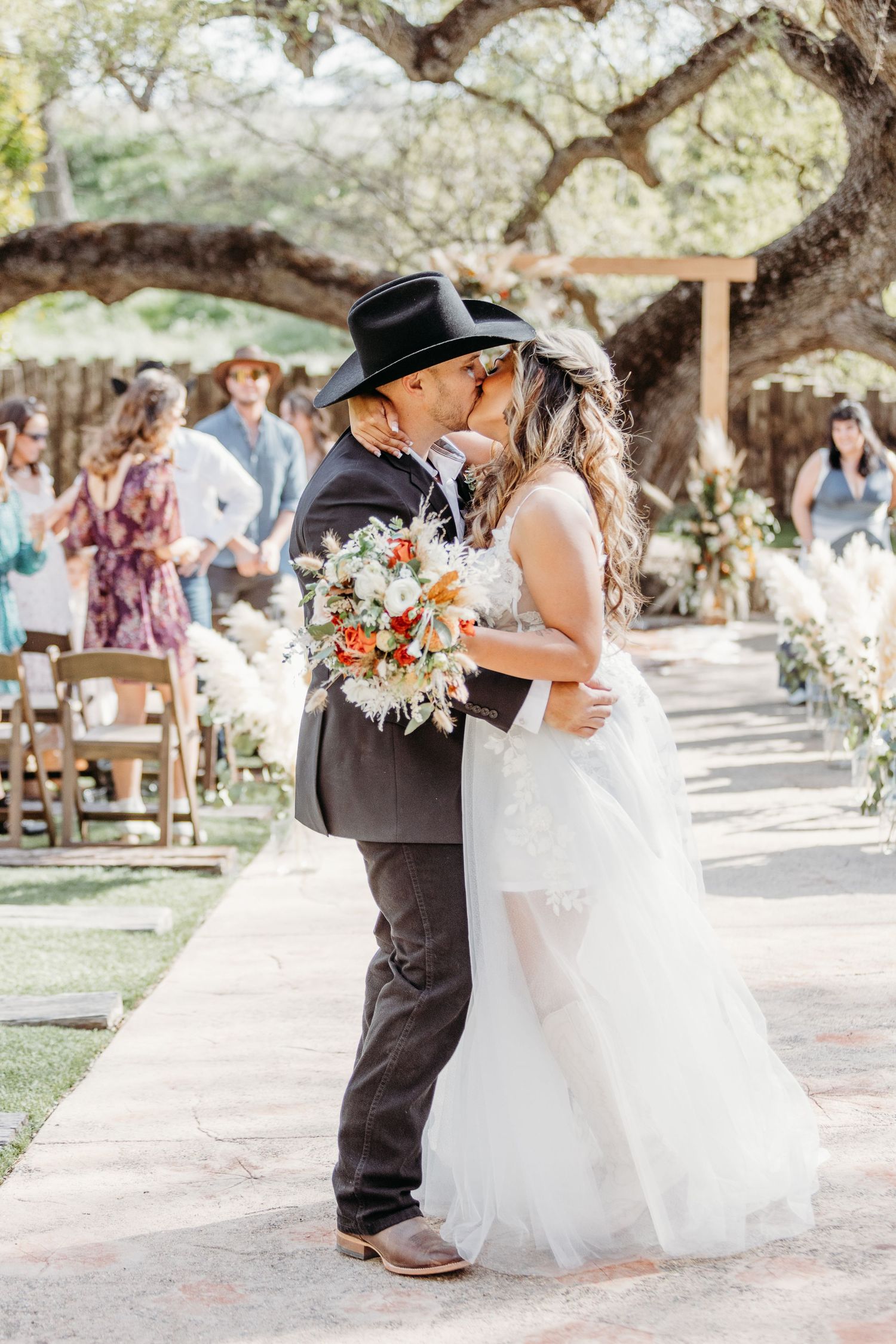 A bride and groom share a tender kiss during their outdoor garden wedding ceremony under sprawling oak trees.