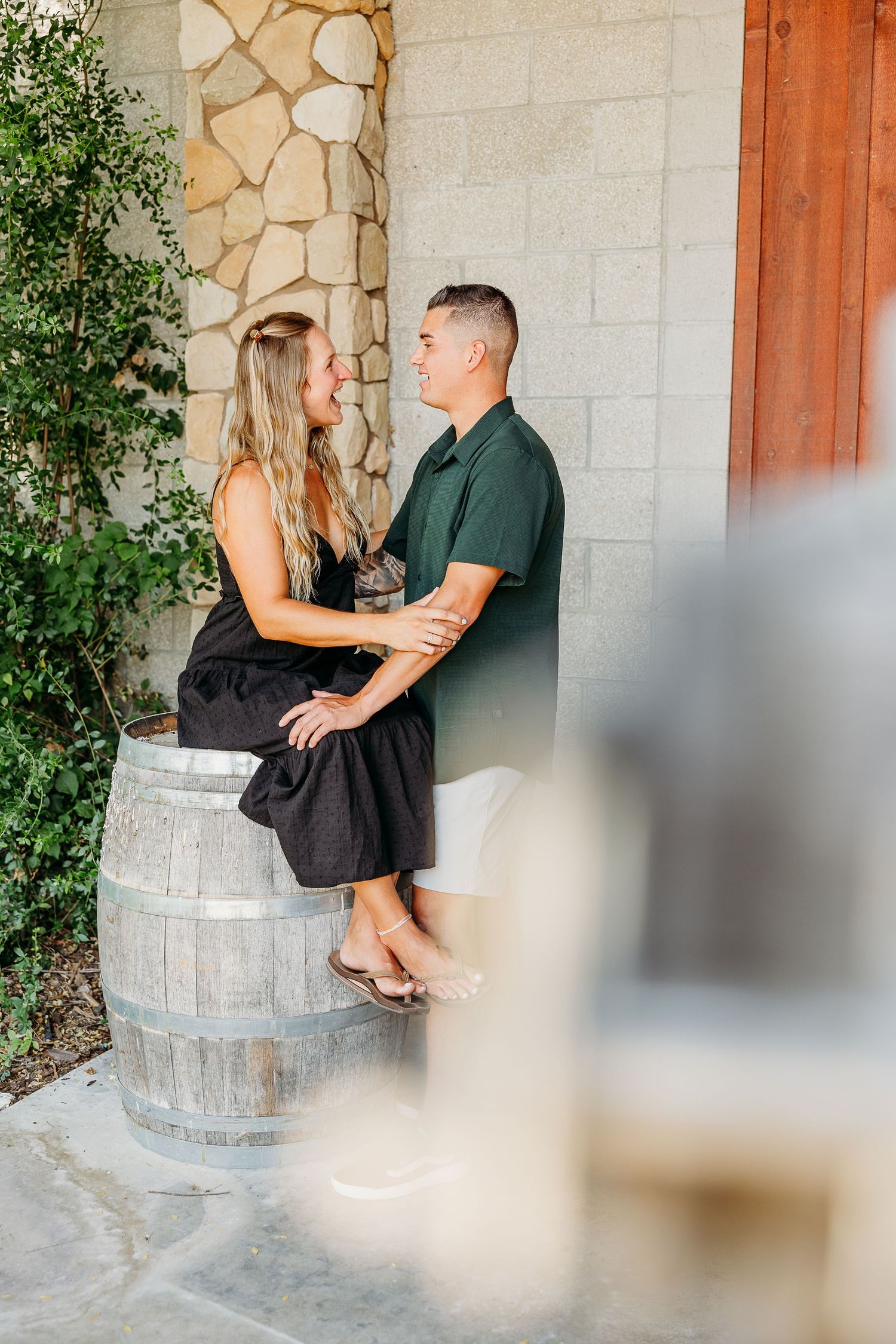 Romantic couple sitting on rustic wine barrels against a stone wall during an engagement photo session.