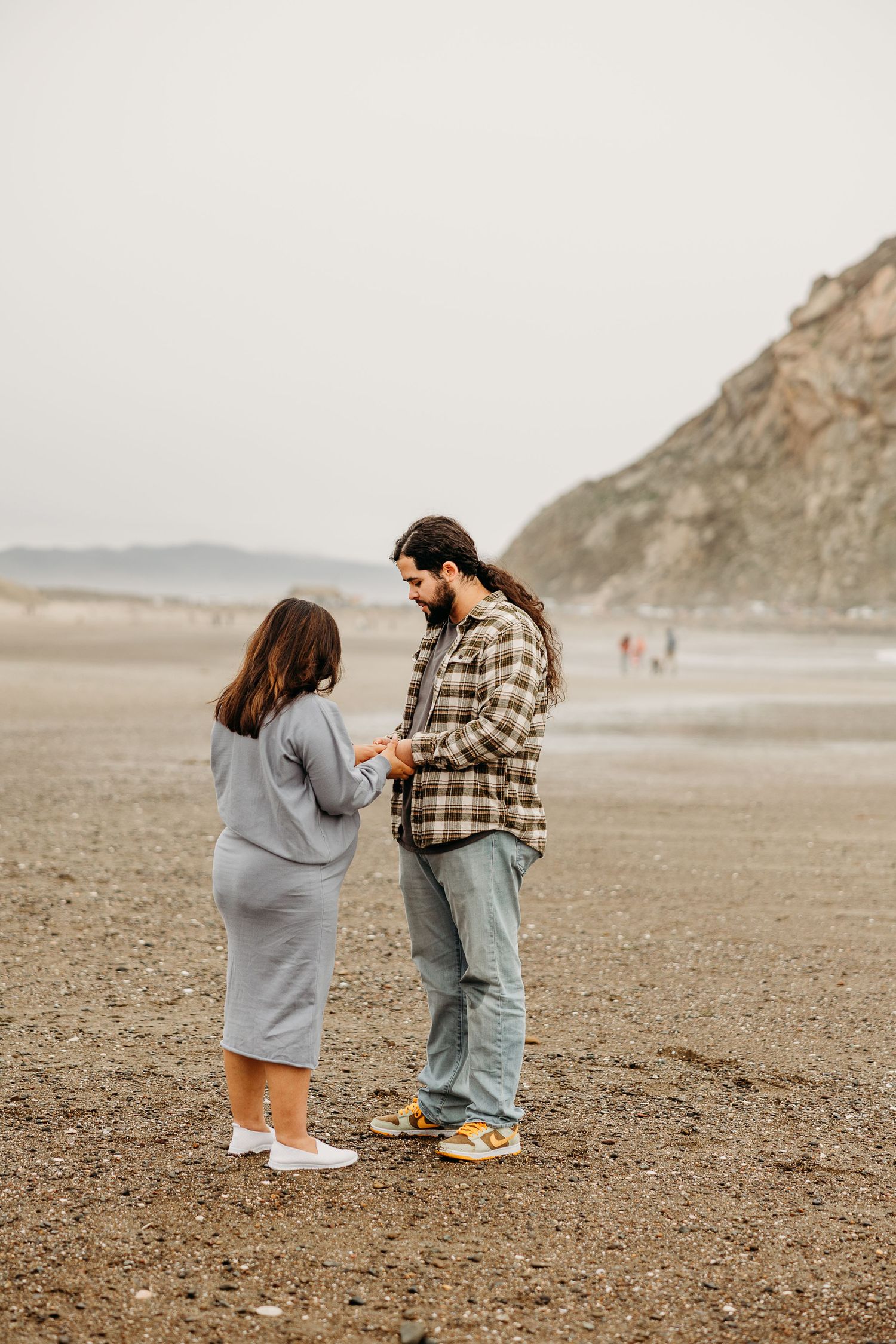 A couple stands together on a foggy beach with cliffs in the background.