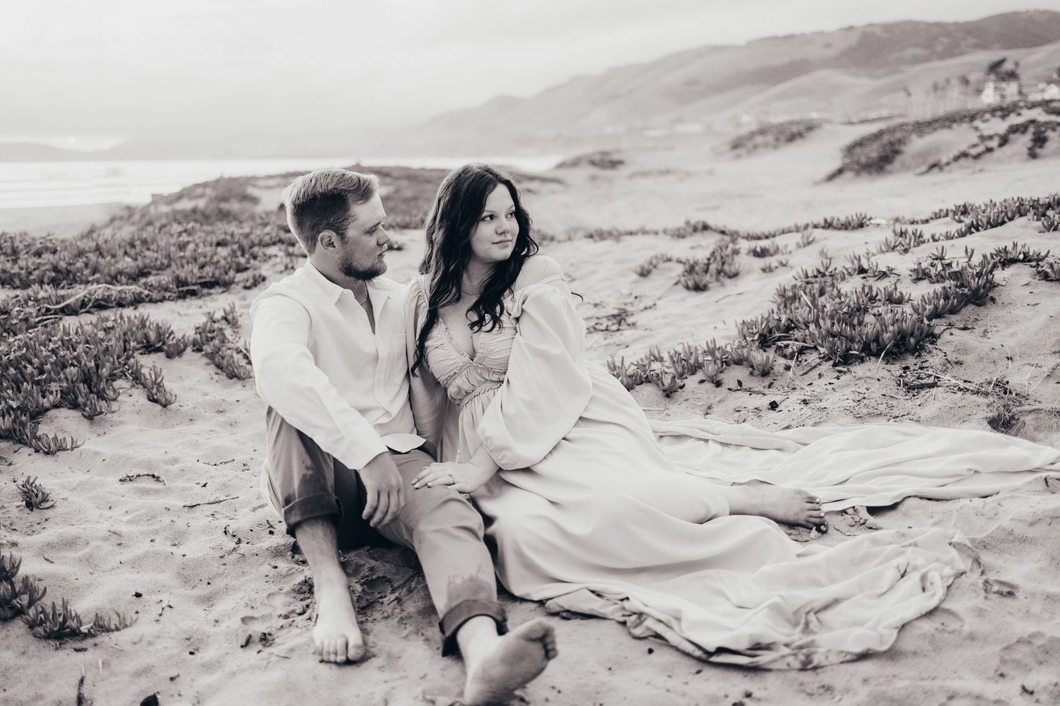 Monochrome capture of couple sitting on coastal dunes with mountains in background.