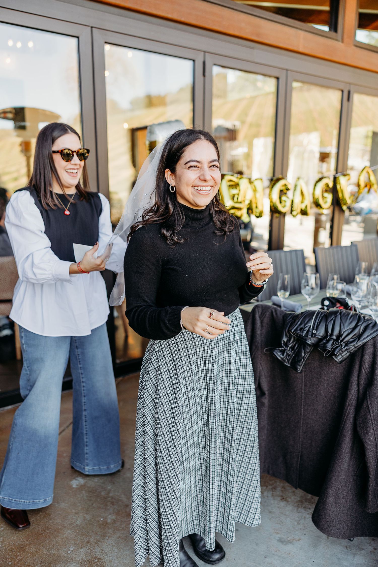 Two women celebrating at an engagement party with gold balloon letters visible in the background.