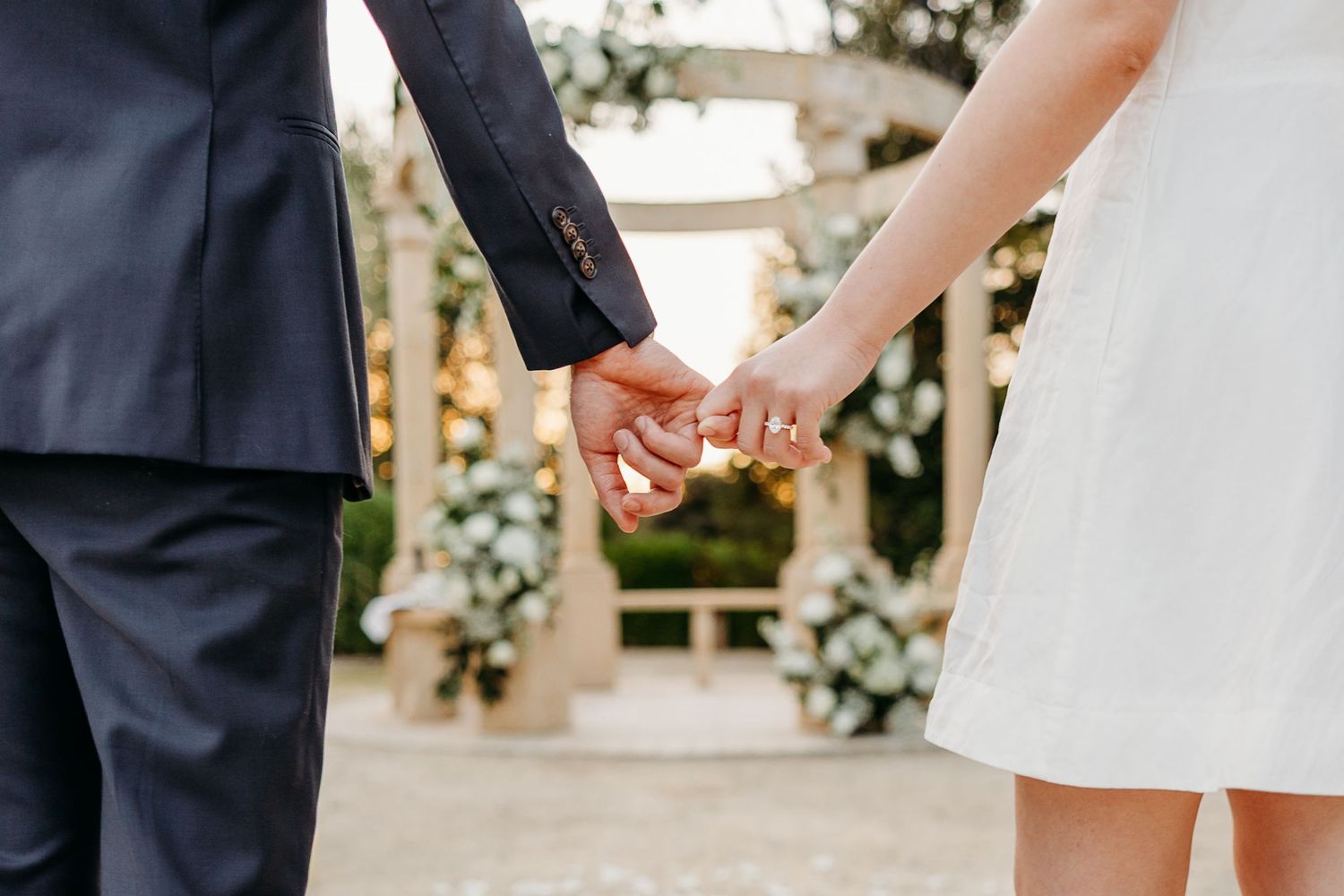 Close-up view of hands holding during an outdoor wedding ceremony with floral decorations in background.