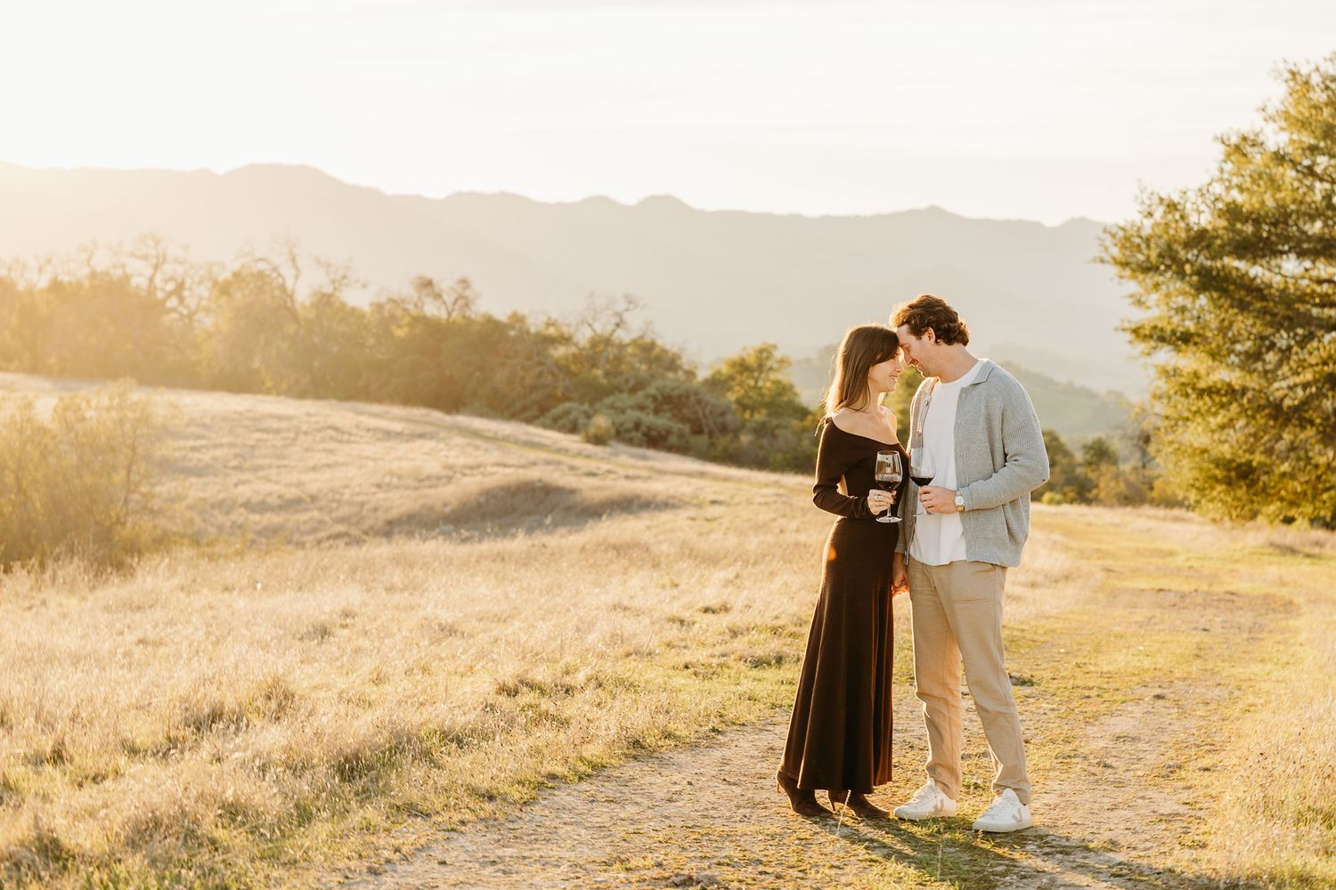 Couple shares intimate moment on hilltop trail during golden hour with mountain backdrop.