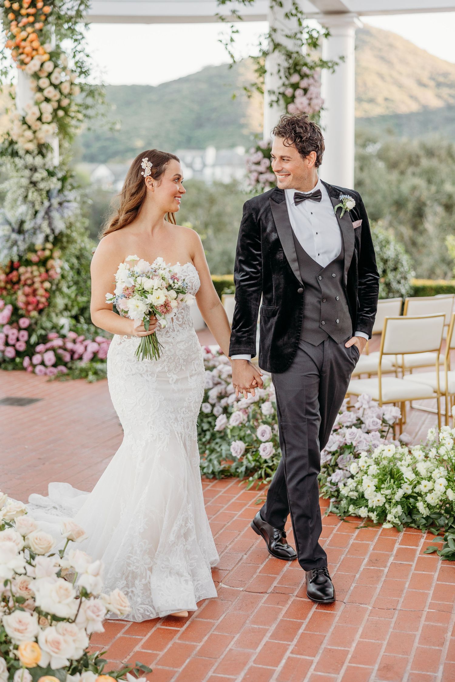 Newlyweds share a romantic moment on a terrace with floral arrangements and mountain views in the background.
