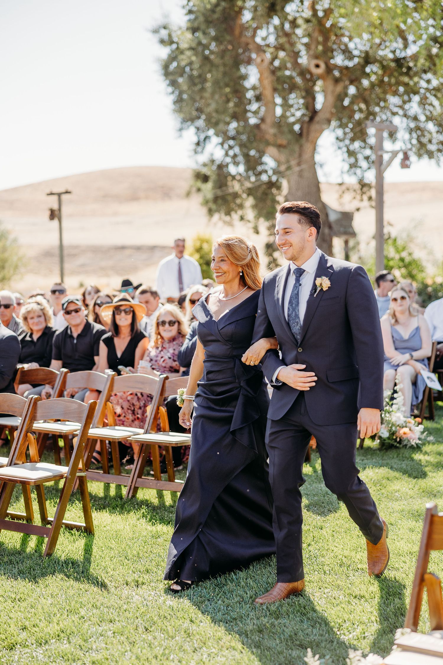 Wedding guests seated on wooden chairs at an outdoor ceremony while a couple embraces under oak trees.