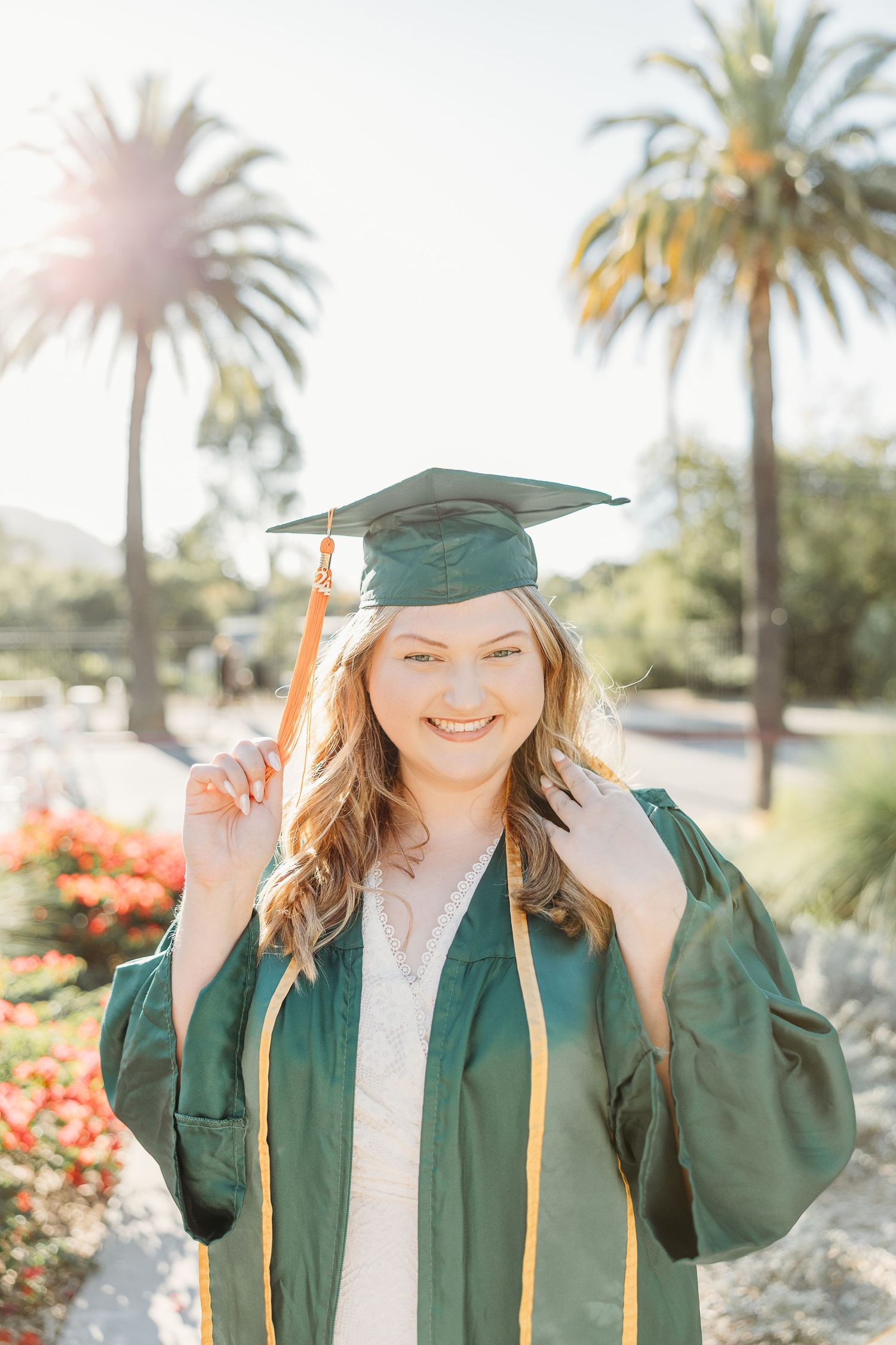 Graduate in green cap and gown smiles brightly in front of palm trees on a sunny day.