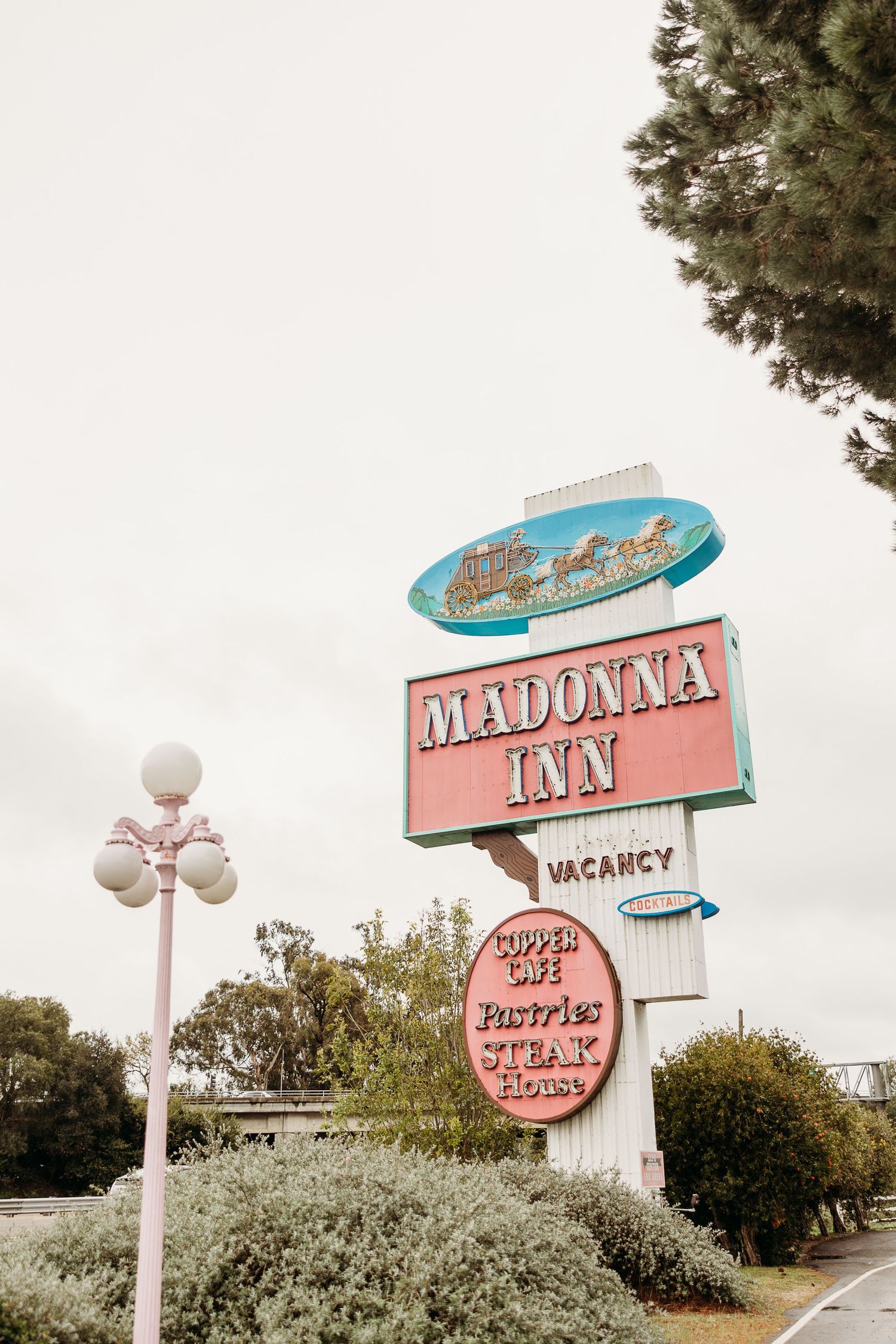 Vintage pink and turquoise Madonna Inn motel sign with classic mid-century design against cloudy sky and landscaping.