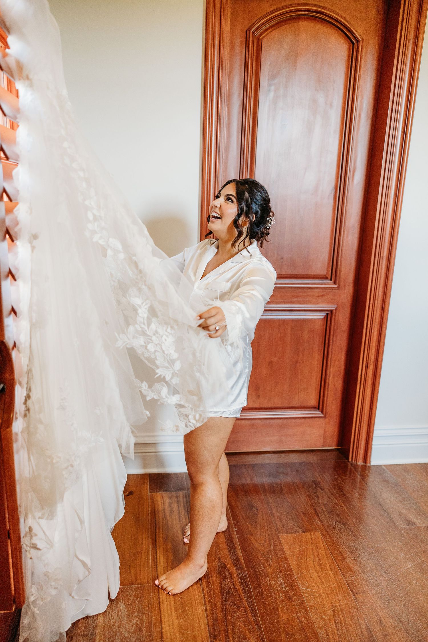 A joyful moment as someone in a white robe playfully holds up a flowing lace wedding dress against a wooden door.