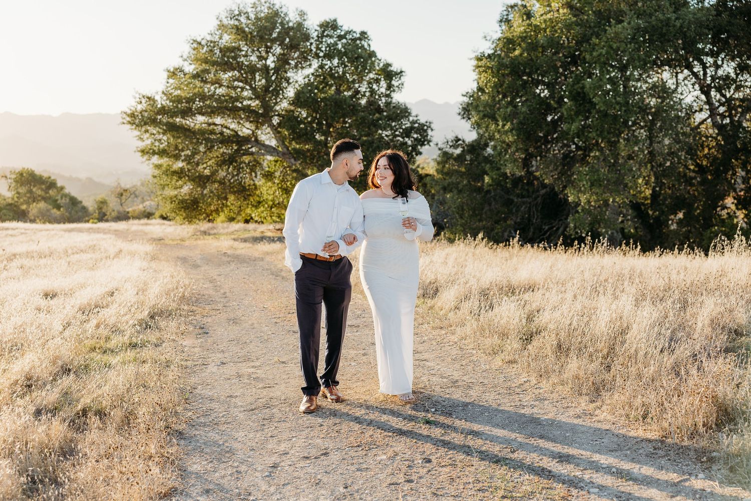 A couple walks together on a sunlit dirt path through golden grass and oak trees in a scenic outdoor setting.