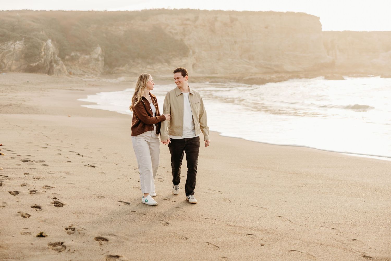 Romantic couple strolling hand in hand along a misty beach at golden hour.