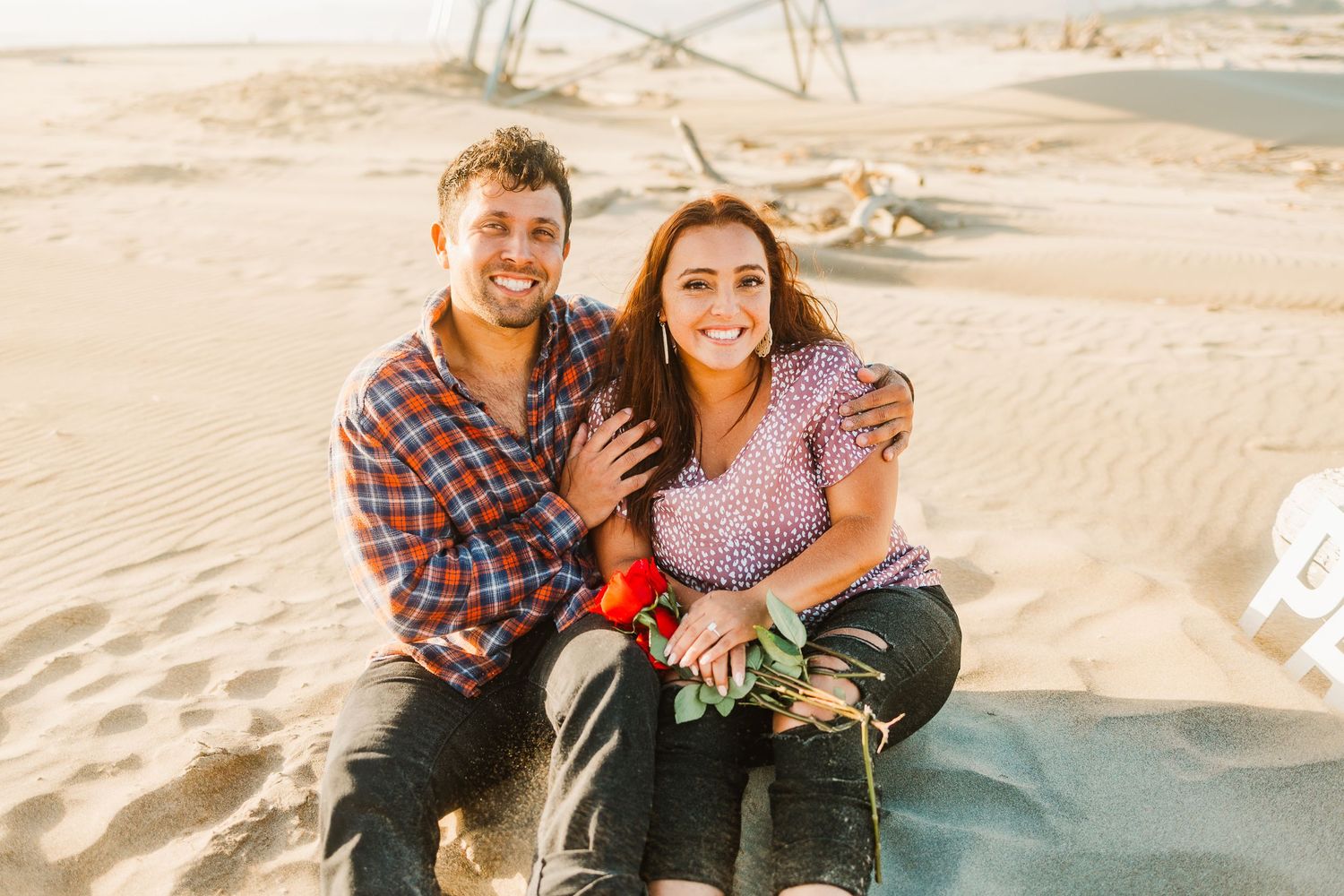A couple sits together on sandy dunes during a beach photoshoot.