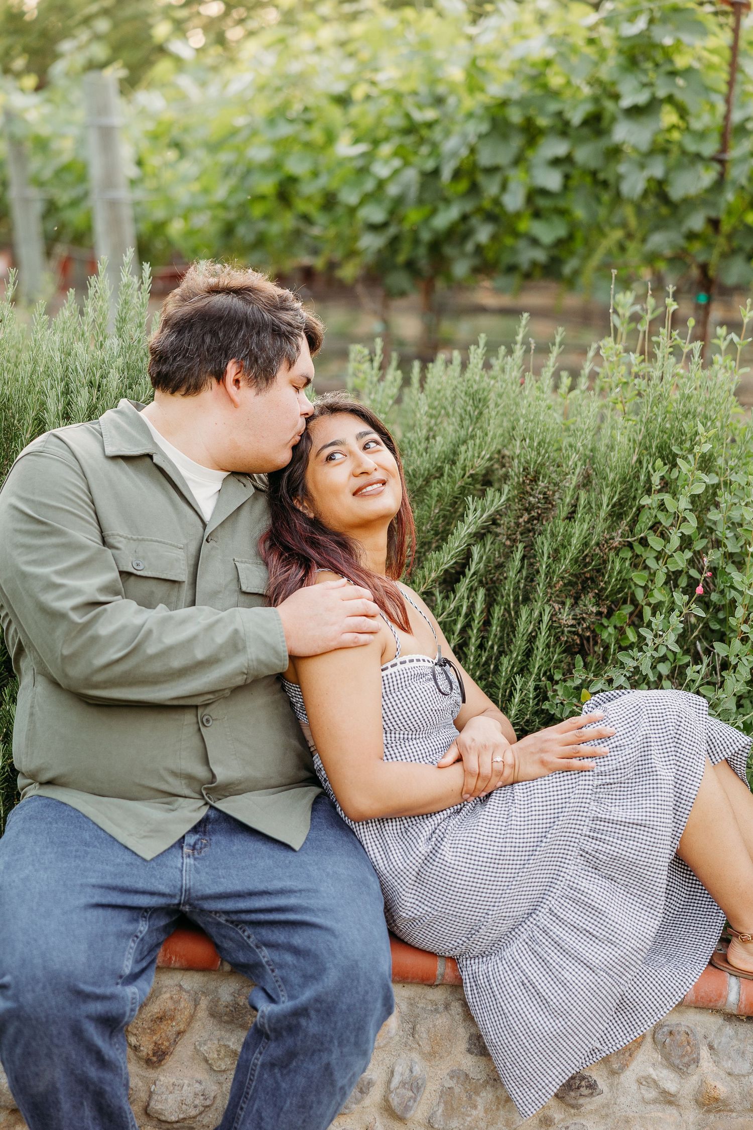 A romantic couple sits together on a stone wall surrounded by lush garden herbs and greenery.