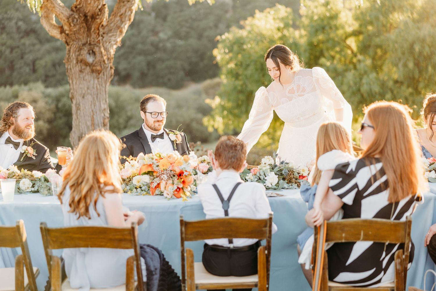 Outdoor wedding reception with table settings and guests enjoying dinner at sunset.