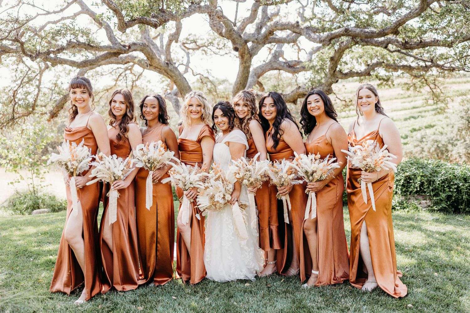 Full bridal party in rust-colored dresses poses outdoors under sprawling oak trees.
