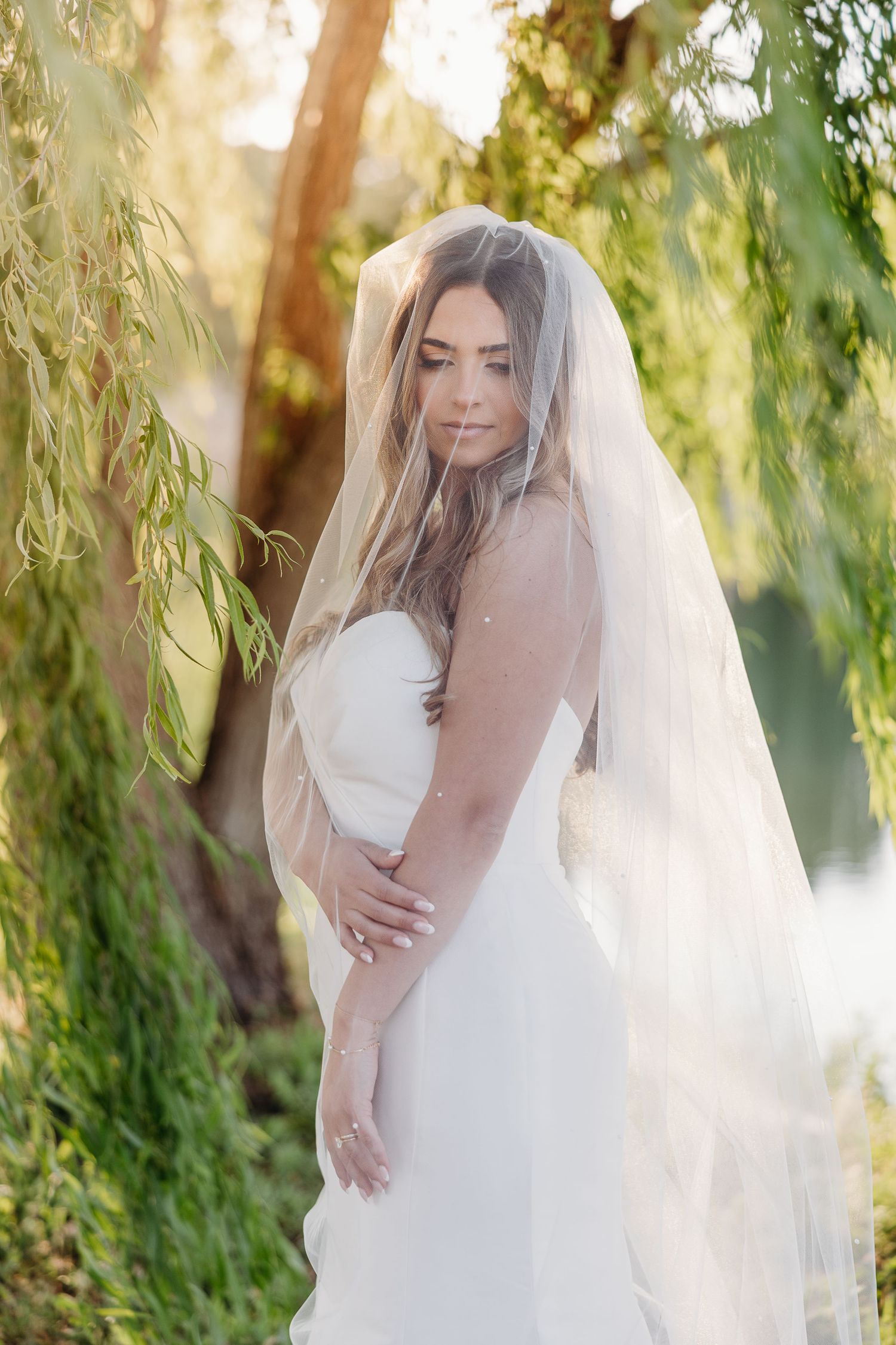 Dreamy bridal portraits under sunlit willow trees showcase flowing white wedding dress and cathedral length veil.