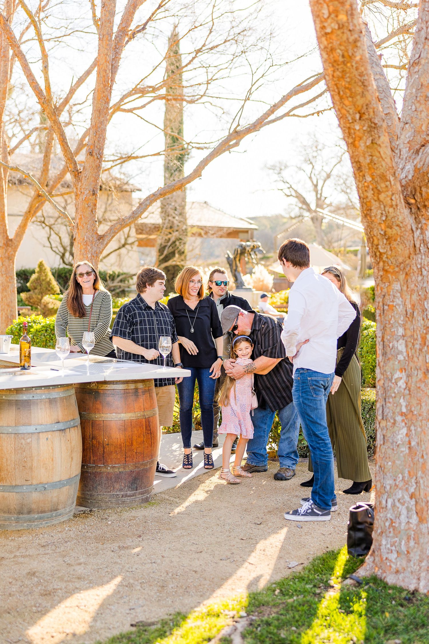 Friends and family gather around wine barrels for a celebration in a sunny outdoor setting.