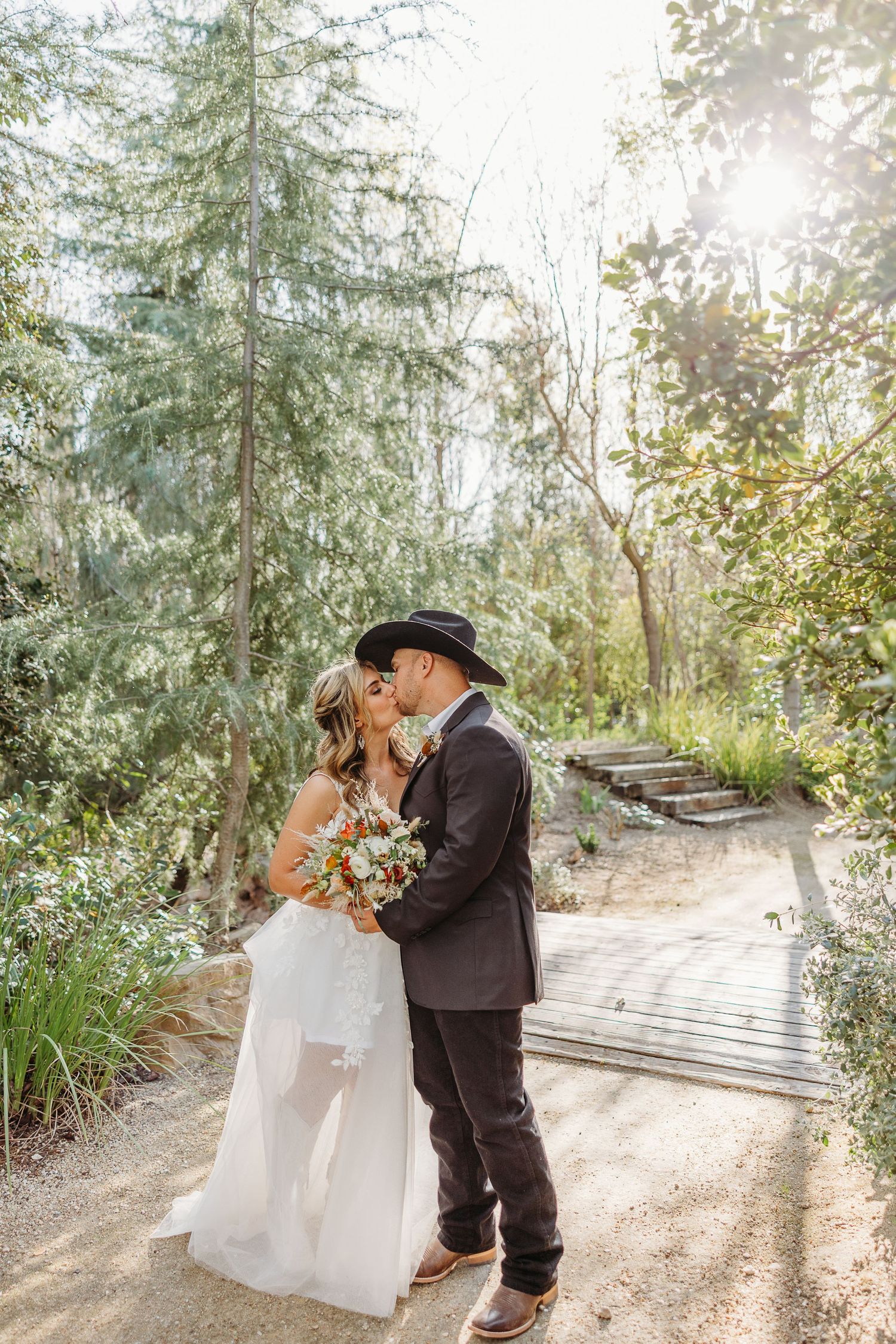 A romantic wedding couple shares a tender moment on a sunlit garden path surrounded by evergreen trees and foliage.