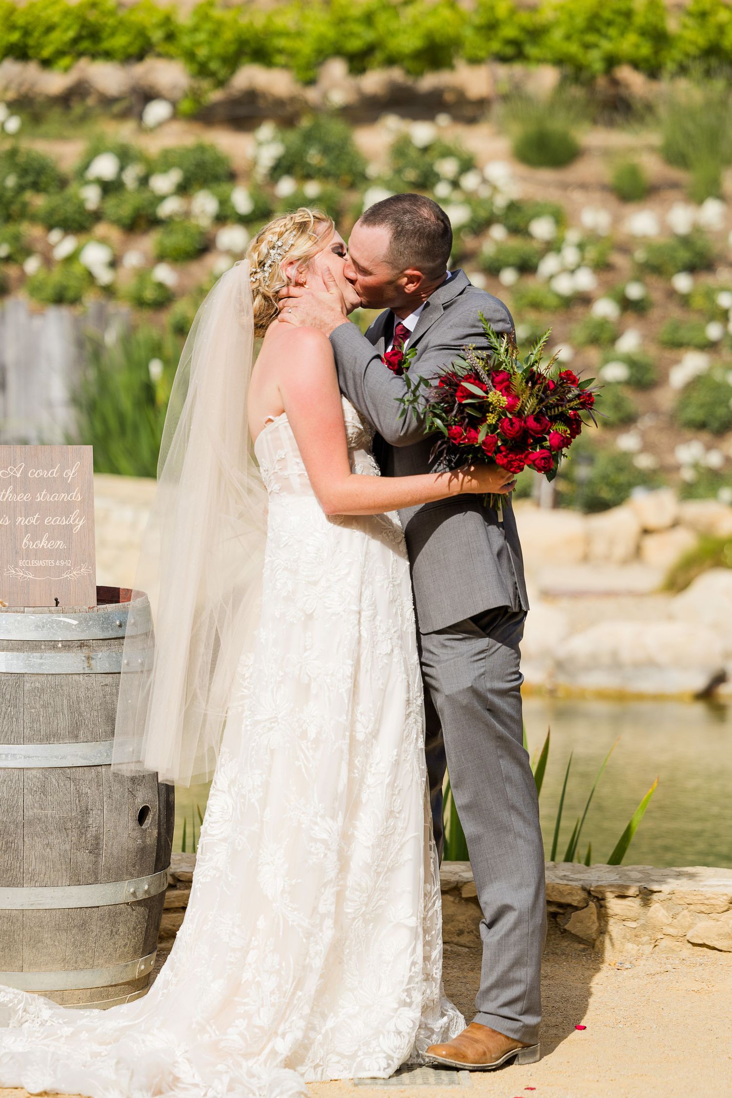 Romantic wedding kiss with vibrant red bouquet and wine barrel decor.