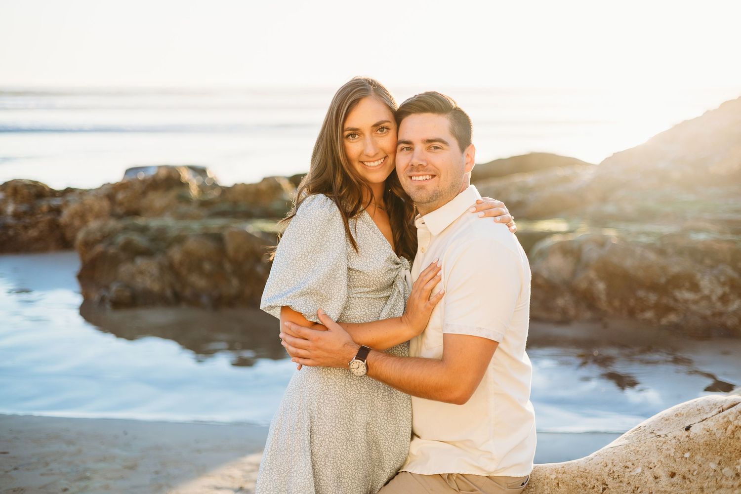 A couple smiles at the camera while embracing on a rocky beach at sunset.