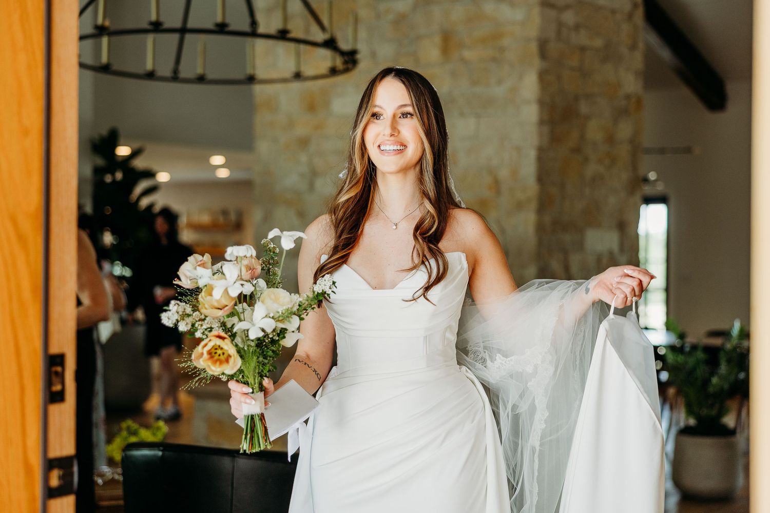 Smiling bride in white gown holding peach and white bouquet standing in elegant venue doorway.