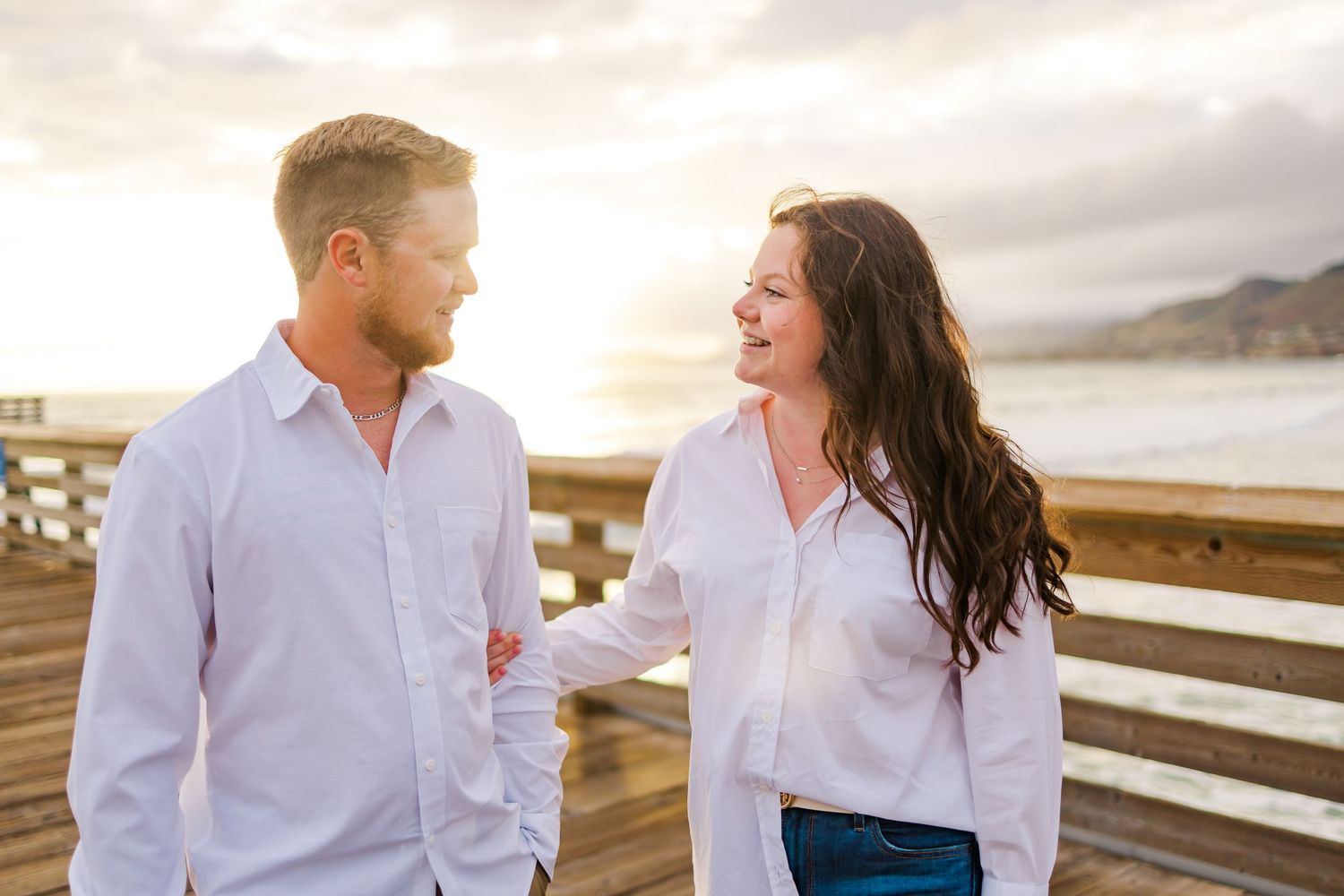 A joyful couple in white shirts sharing a laugh while standing on an oceanside pier.