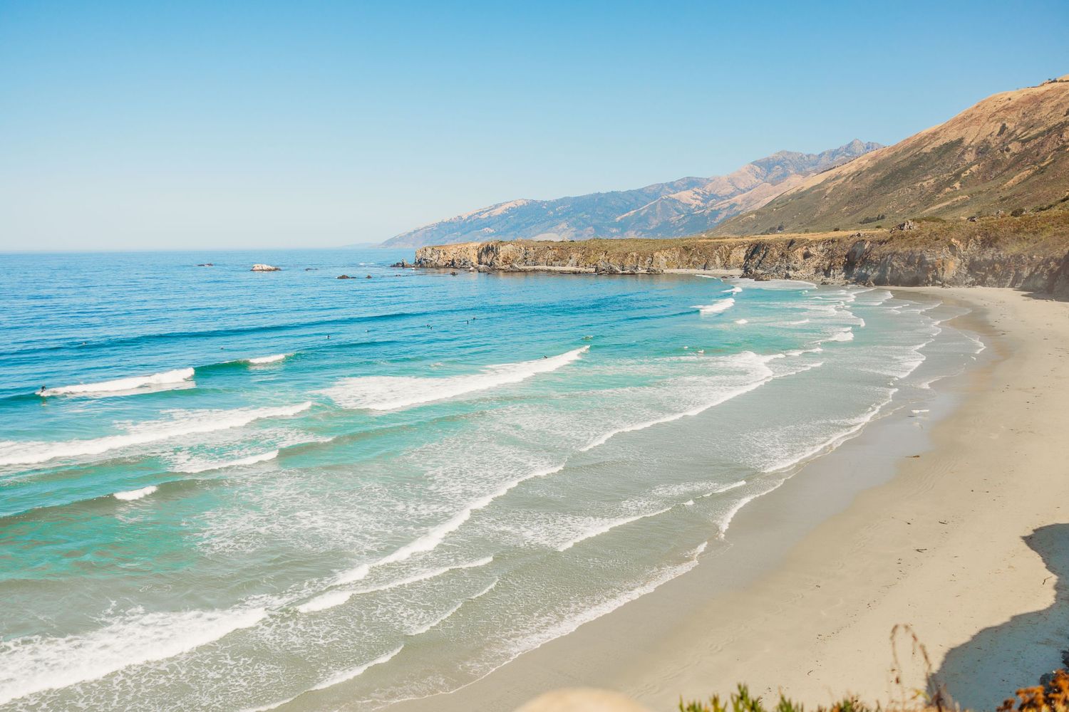 Dramatic coastal landscape showing Big Sur's rugged cliffs and turquoise waves rolling onto sandy beach.
