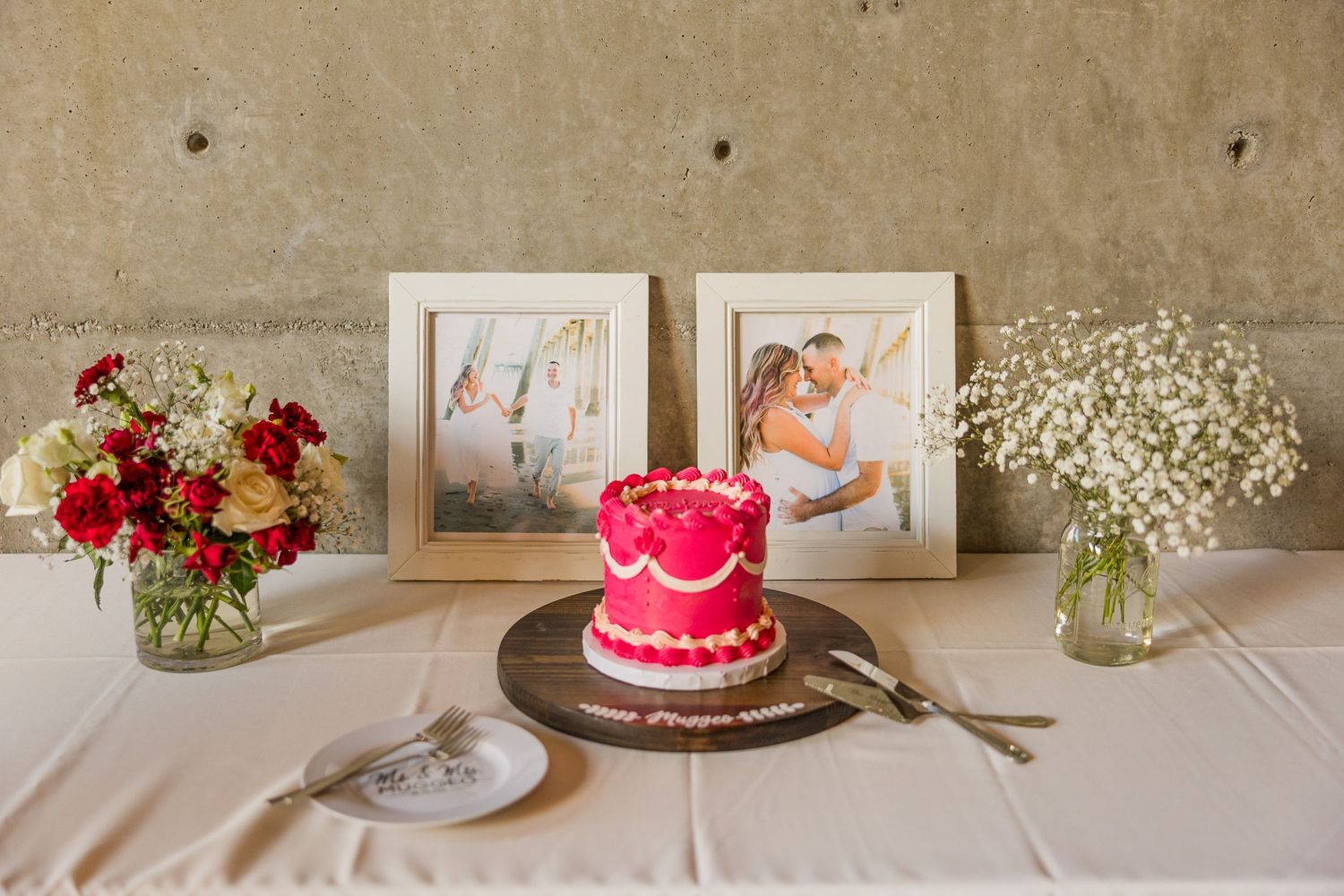Wedding reception table with pink cake, framed photos, and red and white floral arrangements.