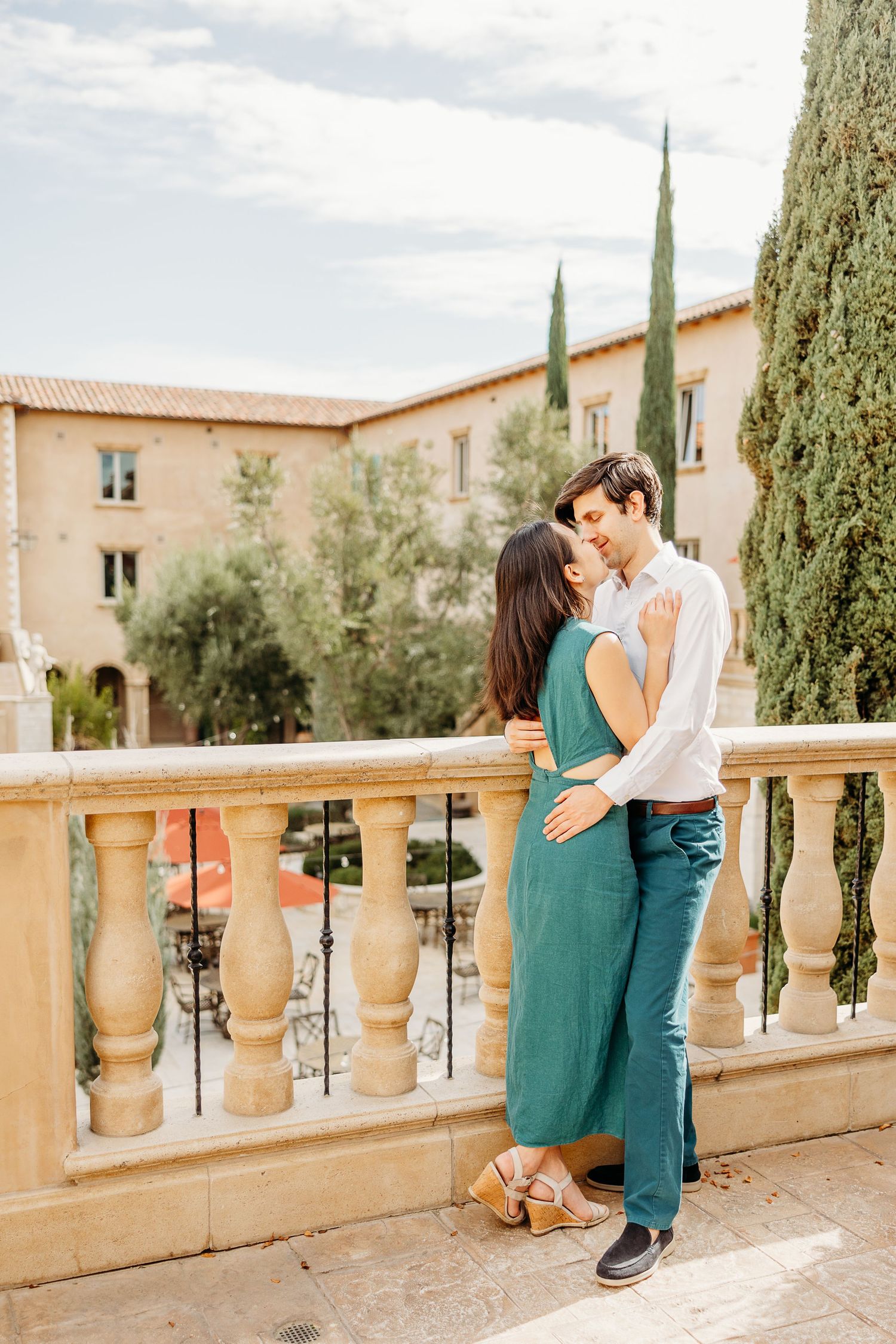 A couple shares romantic kisses on a Mediterranean-style terrace with stone balustrades and cypress trees in the background.